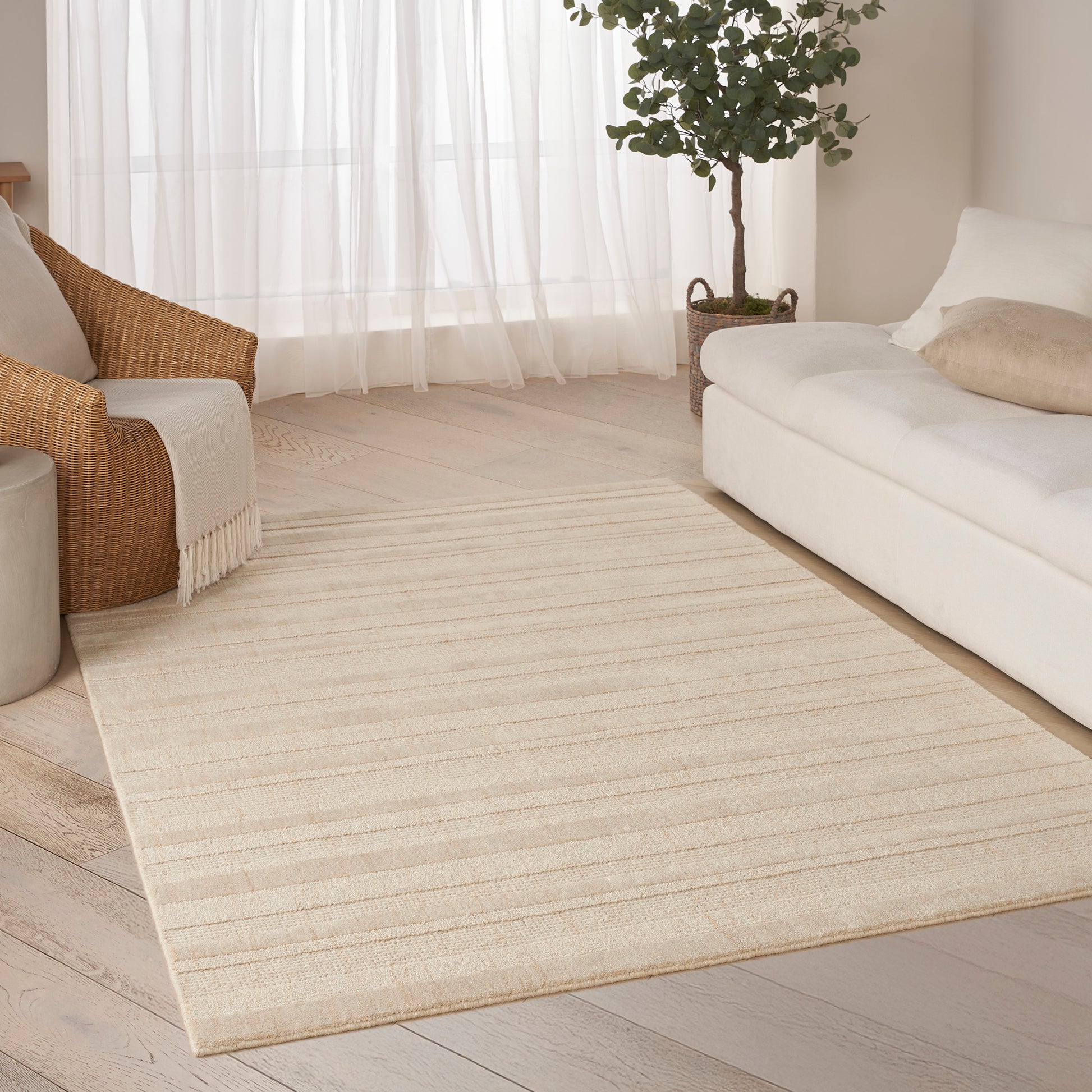 Beige rectangular woven area rug with stripes on light wood floor in a bright living room with cream sofa and wicker chair.