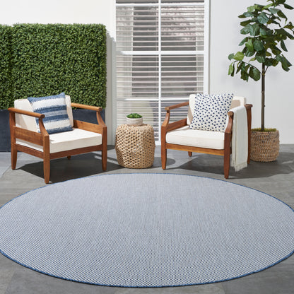 Round blue woven outdoor rug with two wooden armchairs, white cushions, blue/white pillows, and a wicker side table.