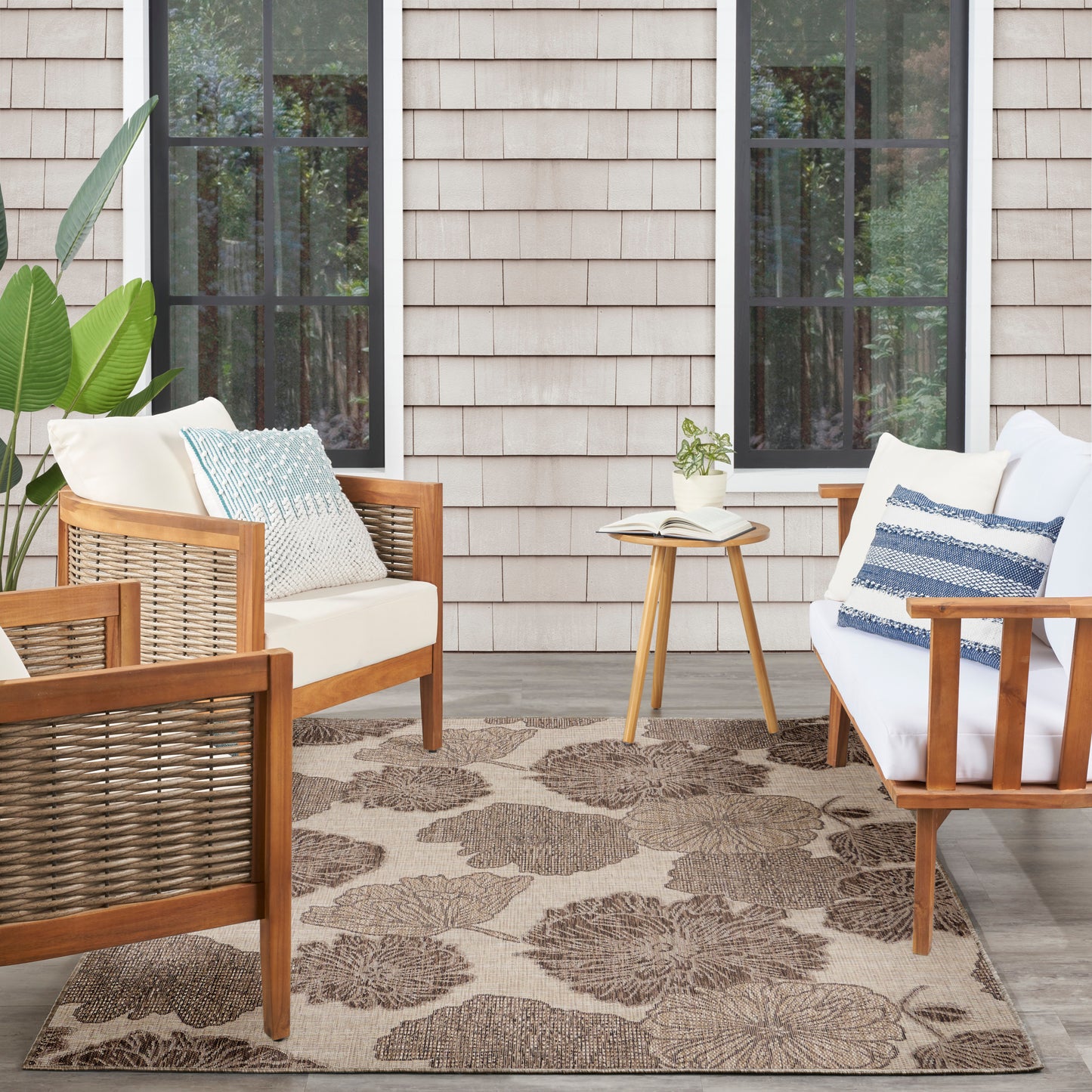 Beige floral outdoor rug with wooden-framed seating featuring cream cushions and blue pillows; small side table with a book.