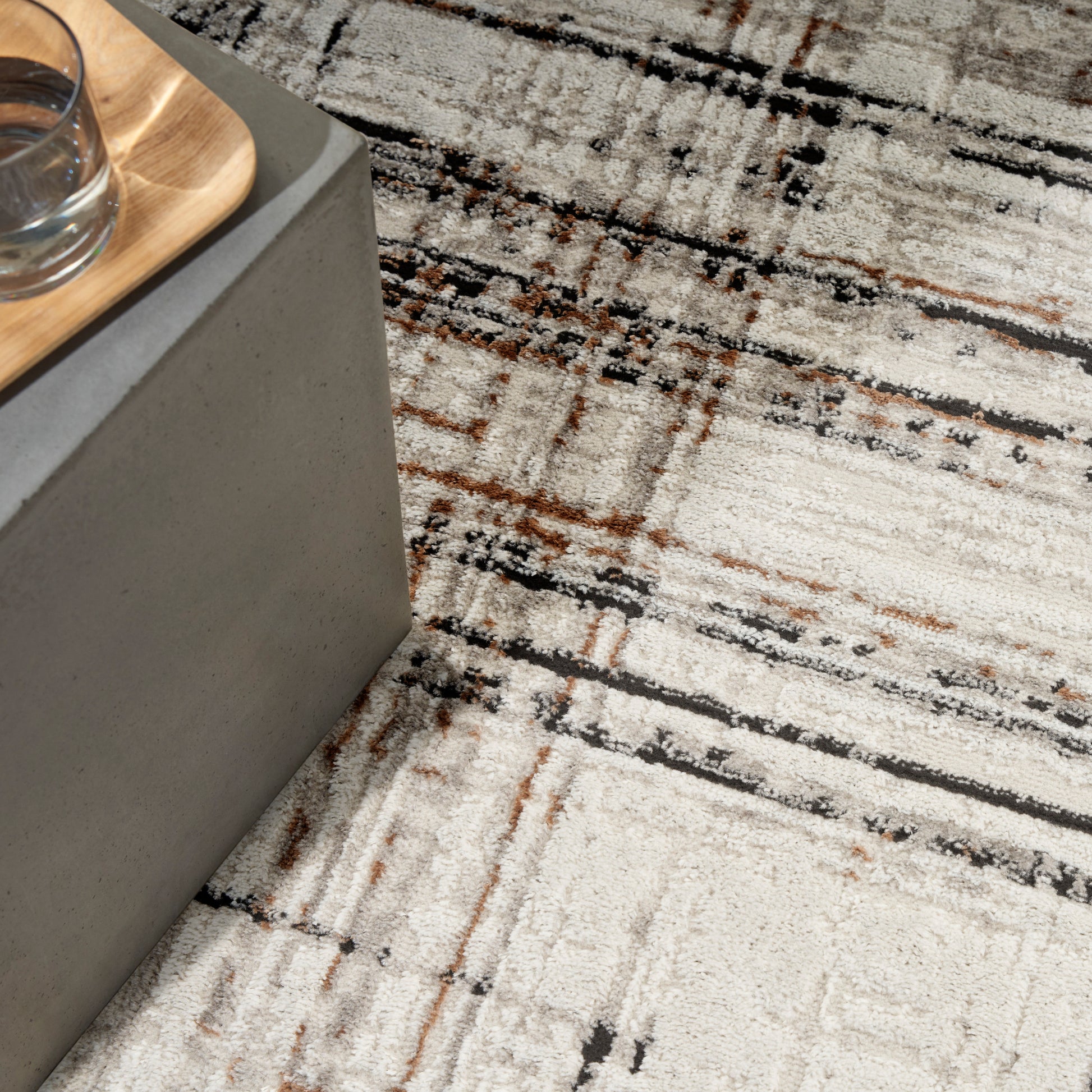 Beige textured rug with black and rust accents, under a gray concrete-block side table with a light wooden tray and glass.