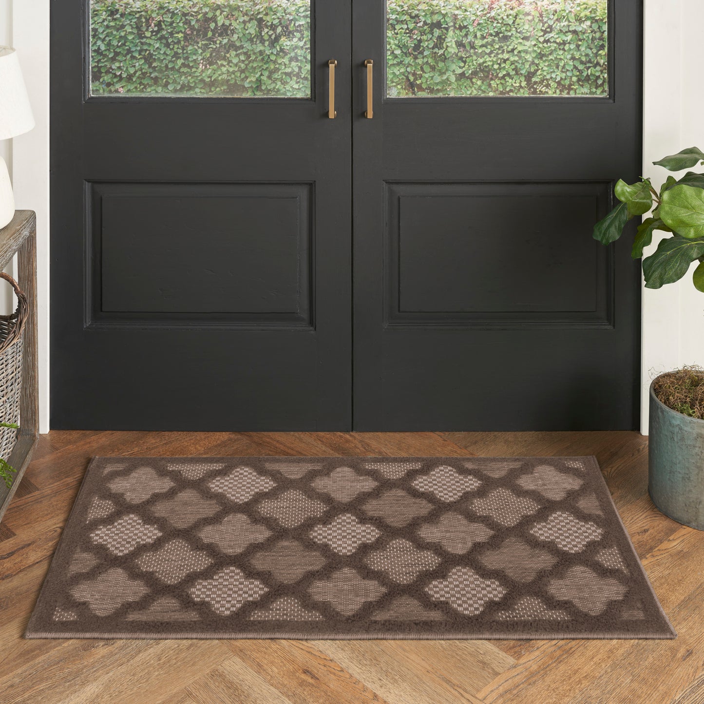 Rectangular doormat with brown border and taupe lattice quatrefoil pattern, placed at a front entry by black double doors.