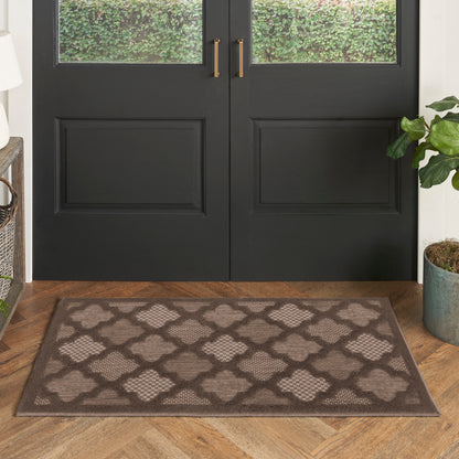 Rectangular doormat with brown border and taupe lattice quatrefoil pattern, placed at a front entry by black double doors.