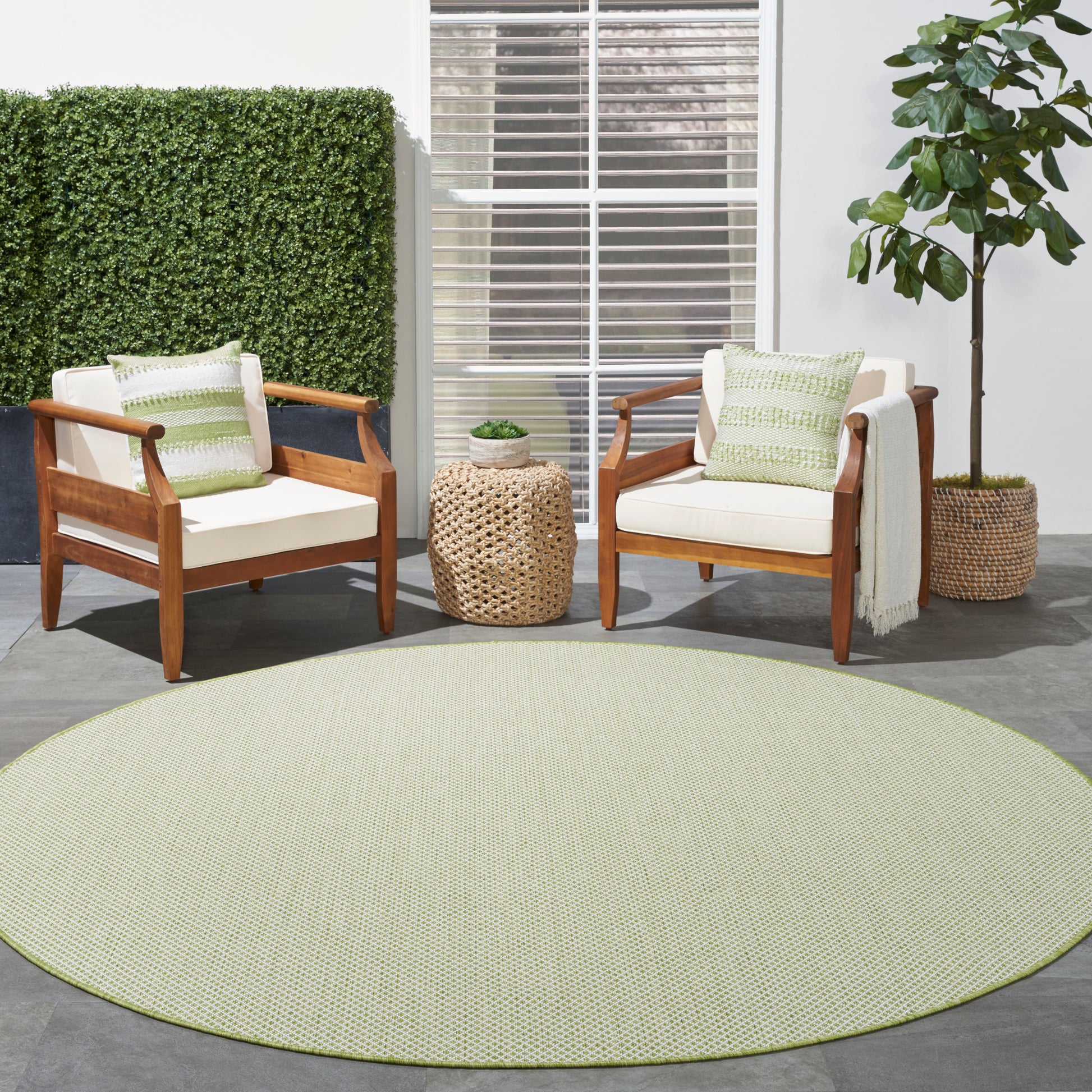 Two wooden armchairs with cream cushions and green pillows, round pale-green rug, woven rattan side table, potted plant.