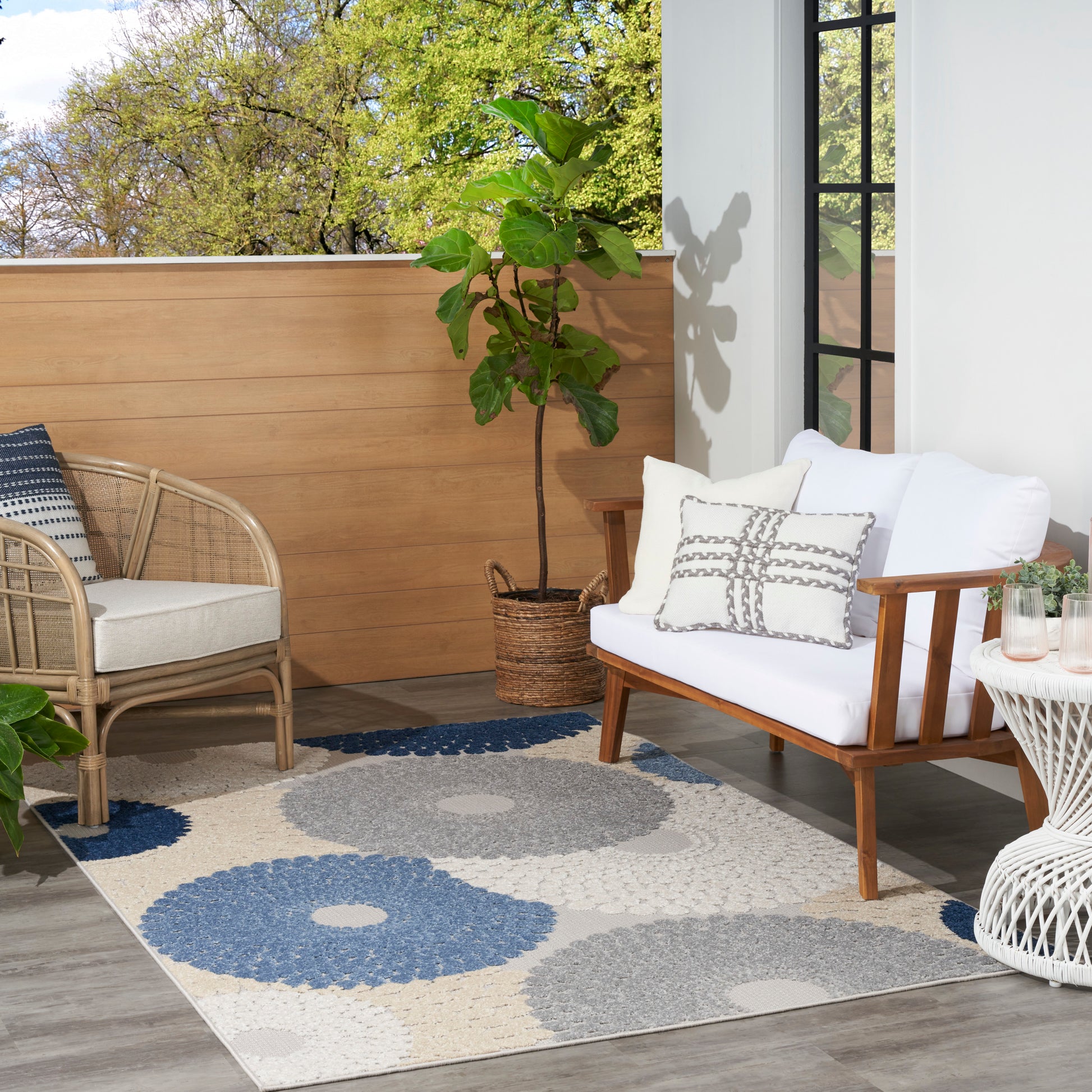 Outdoor balcony arrangement with a blue-gray rectangular rug, white-cushion wood-frame sofa, wicker chair, white side table and plant.