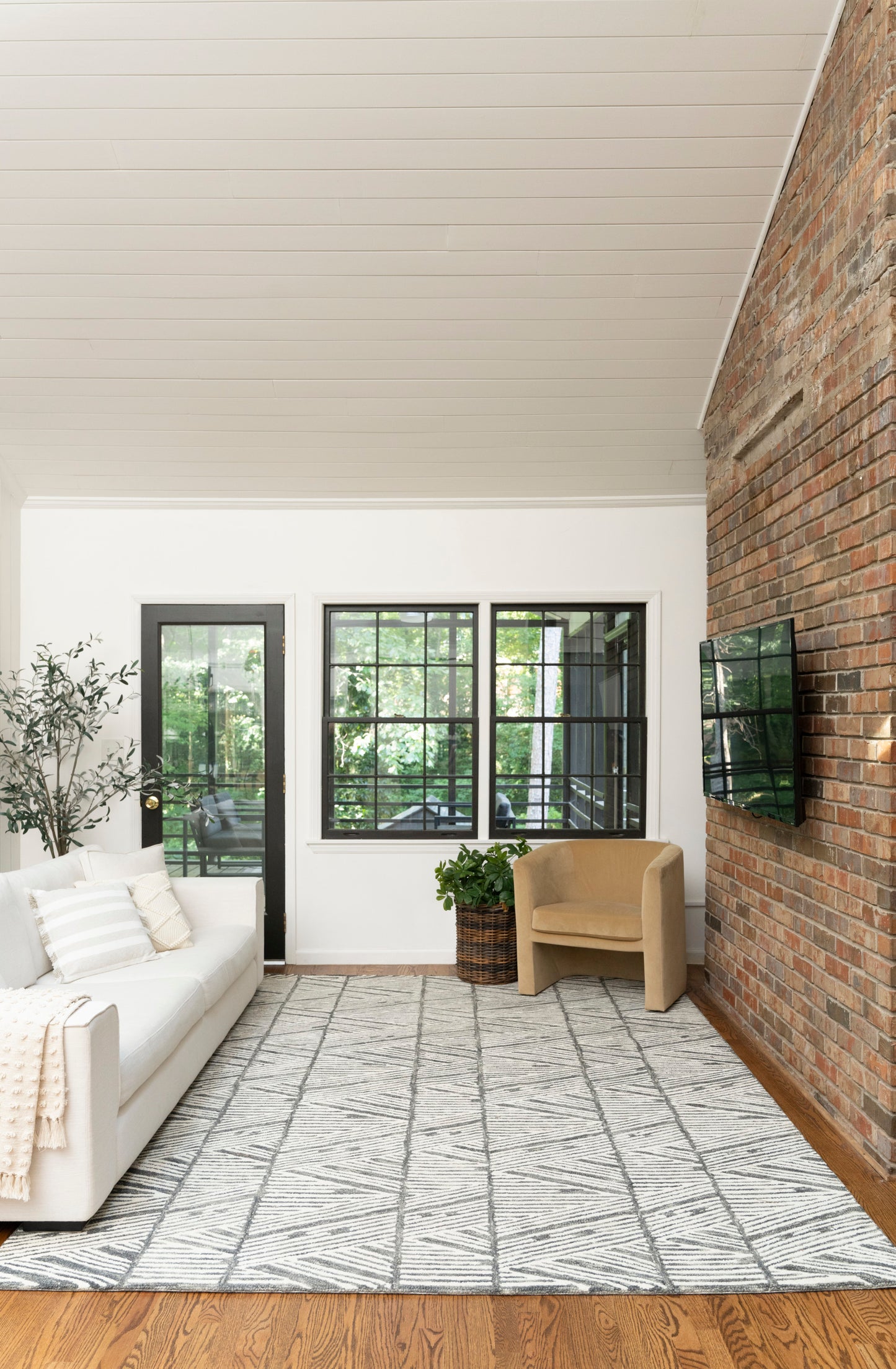 Bright living room with white sofa, beige armchair, black-framed windows and door, brick accent wall, geometric gray rug.