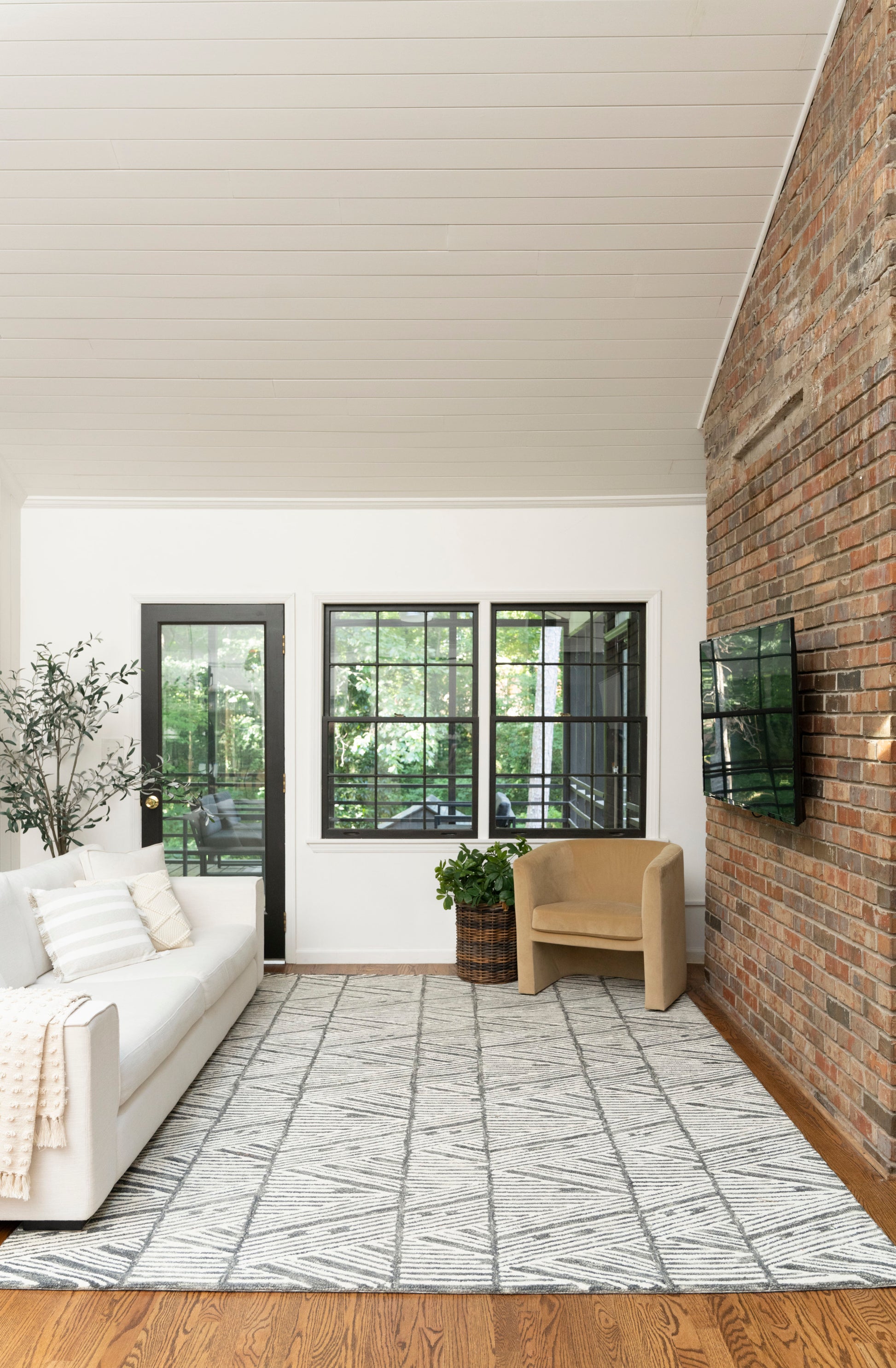 Bright living room with white sofa, beige armchair, black-framed windows and door, brick accent wall, geometric gray rug.
