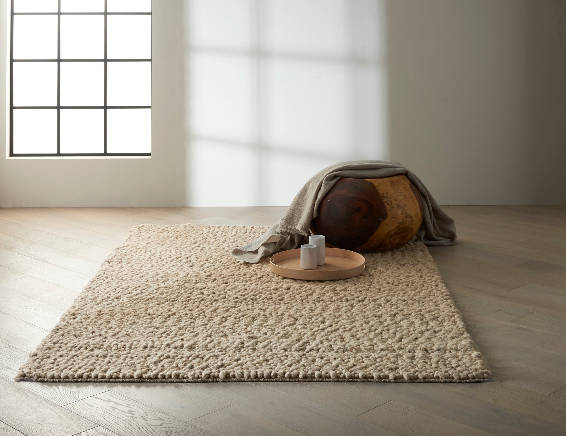 Beige shag rug on light wood floor; round wooden ottoman draped with a gray blanket, tray with two white mugs.