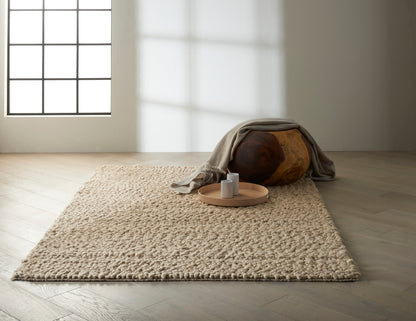 Beige shag rug on light wood floor; round wooden ottoman draped with a gray blanket, tray with two white mugs.