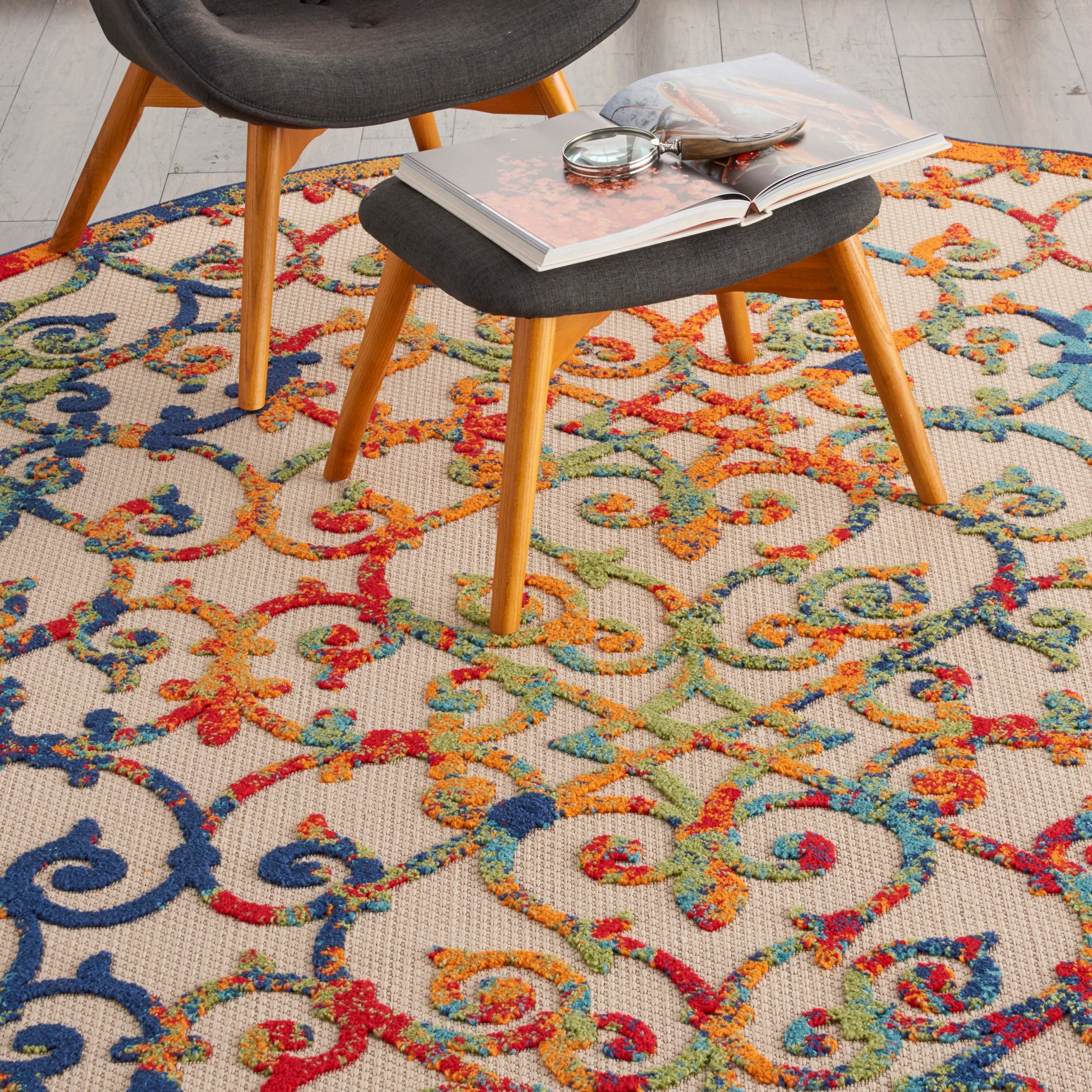 Mid-century modern chairs with light wood legs and dark gray cushions on a vivid multicolor rug; open book on a small stool.