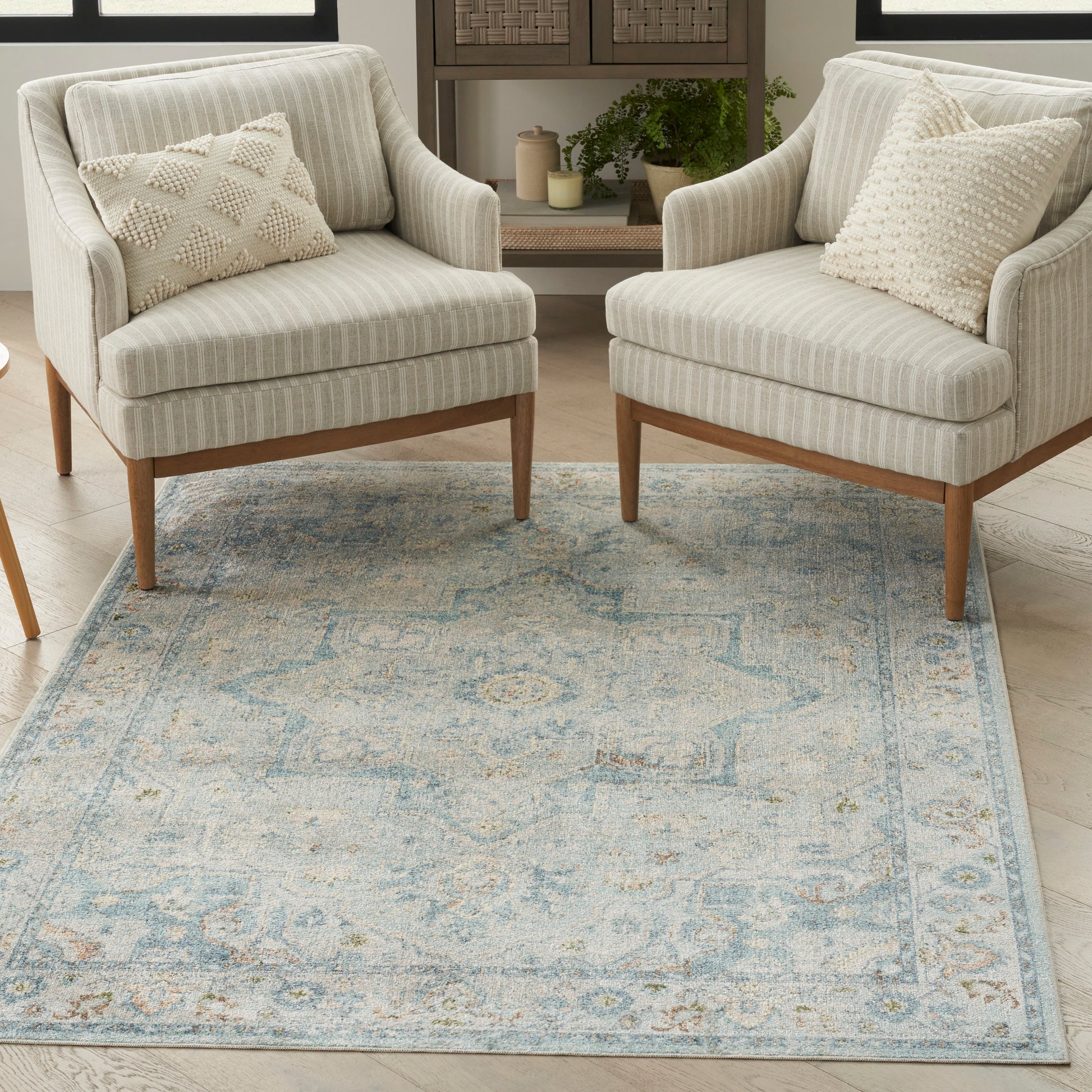 Two light gray striped armchairs with wooden legs flank a blue patterned rug in a cozy living room with plants and candles