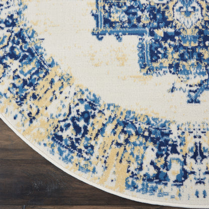 Round area rug with blue, ivory and tan ornate pattern; woven texture with white fringe on dark wood floor.