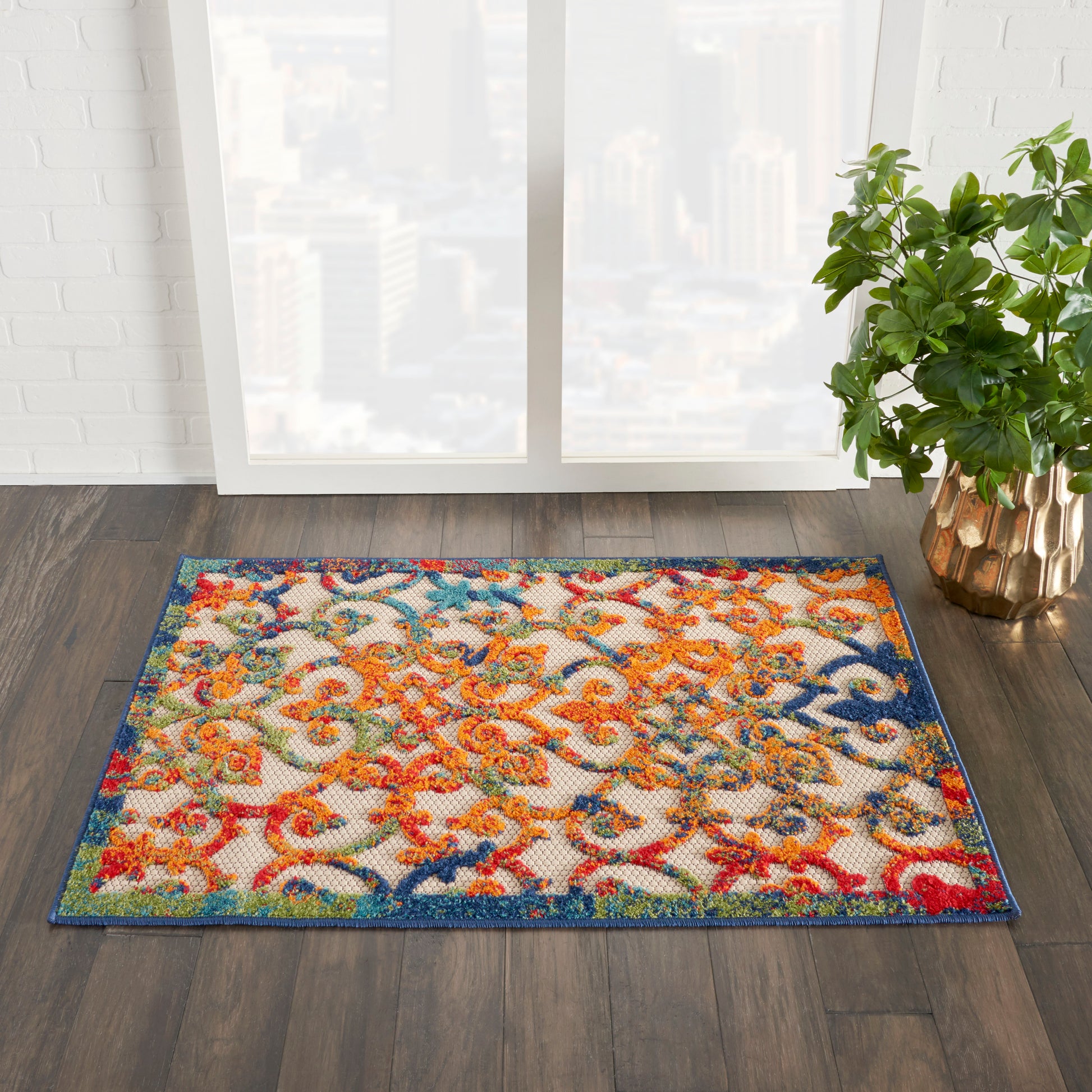 Rectangular woven area rug with a vibrant multicolor floral/scroll pattern and blue border on dark wood floor beside a green potted plant.