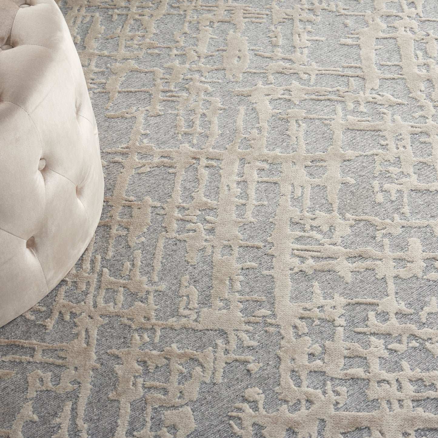 Beige tufted ottoman beside a gray-blue abstract patterned rug.