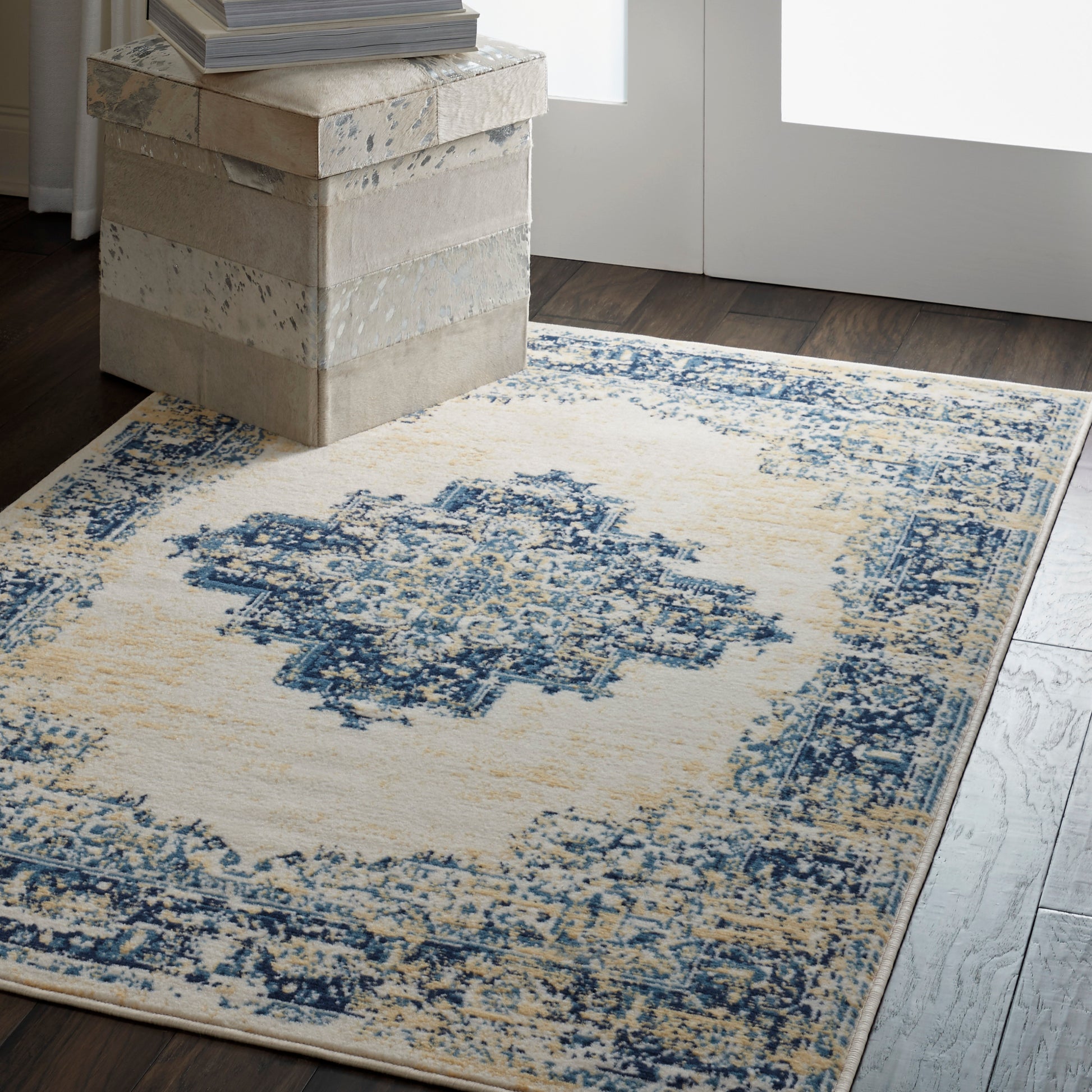Cream distressed rug with blue medallion motif on dark wood floor; beige stackable trunks and books nearby.