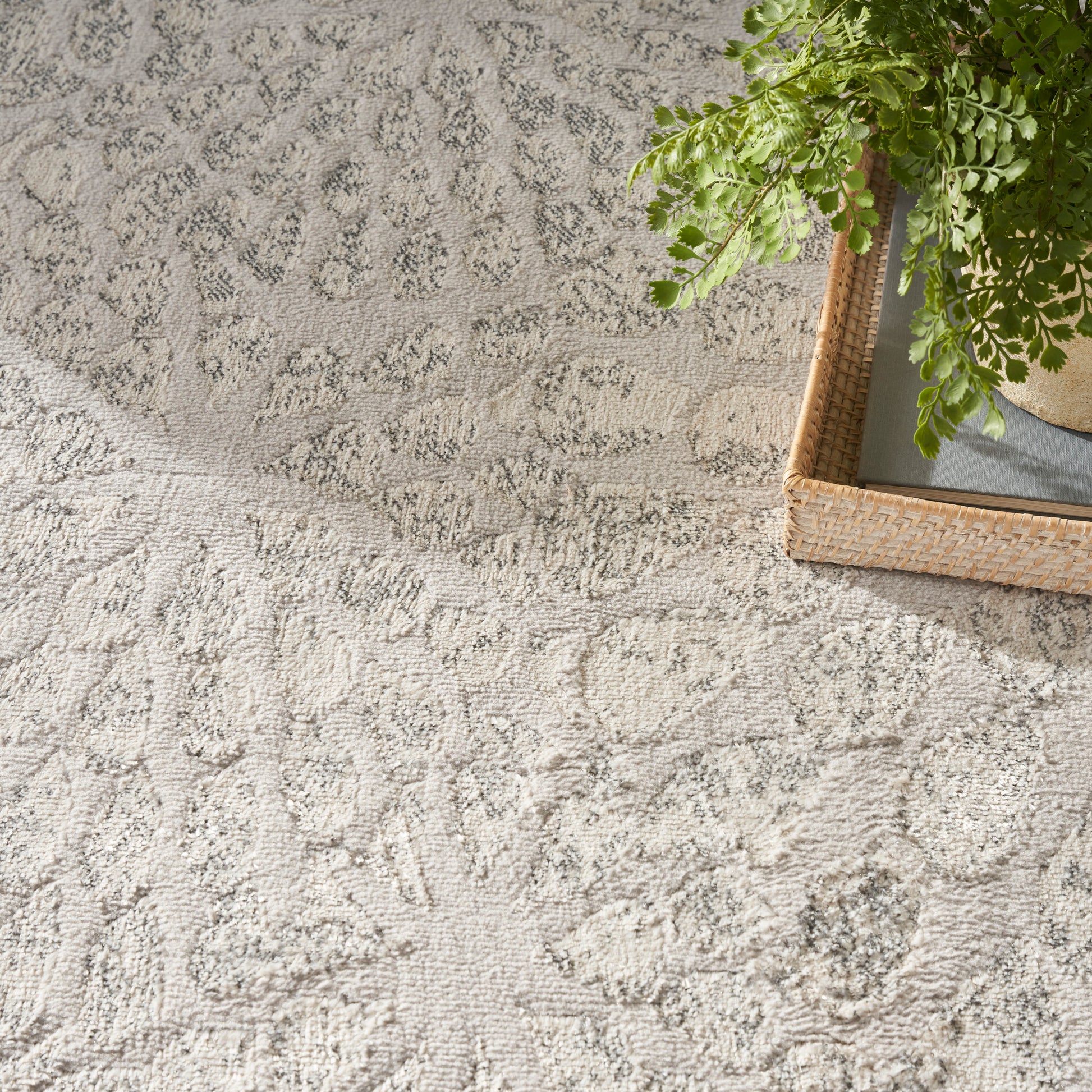 Light gray textured area rug with a subtle floral scroll pattern; corner shows a wicker tray and green fern.