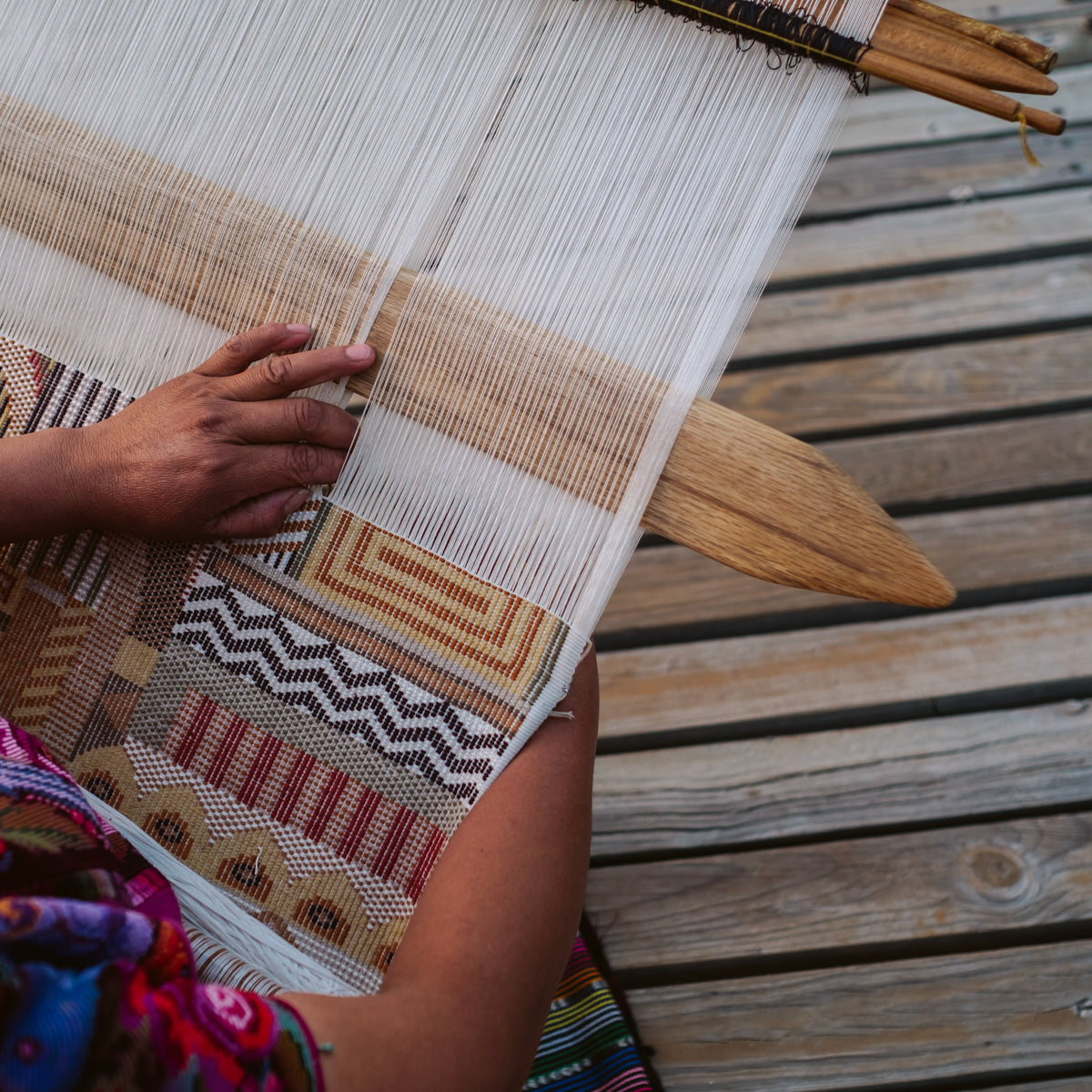 Hands weave white threads through a loom, creating a geometric earthy-toned brocade textile on a wooden loom.