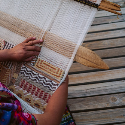 Hands weave white threads through a loom, creating a geometric earthy-toned brocade textile on a wooden loom.