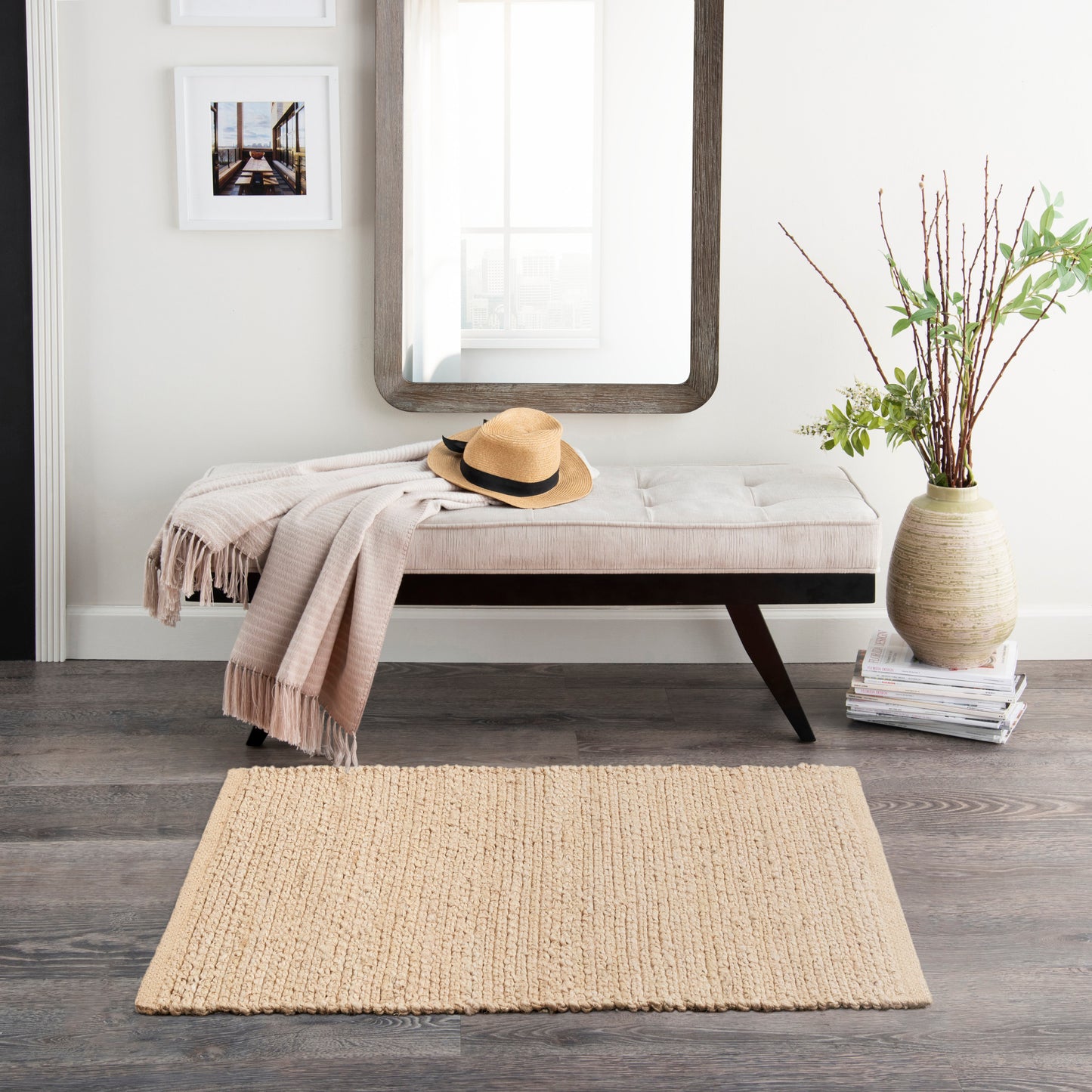 Beige tufted bench with dark legs, throw and straw hat; wood-framed mirror, jute rug, vase with branches.