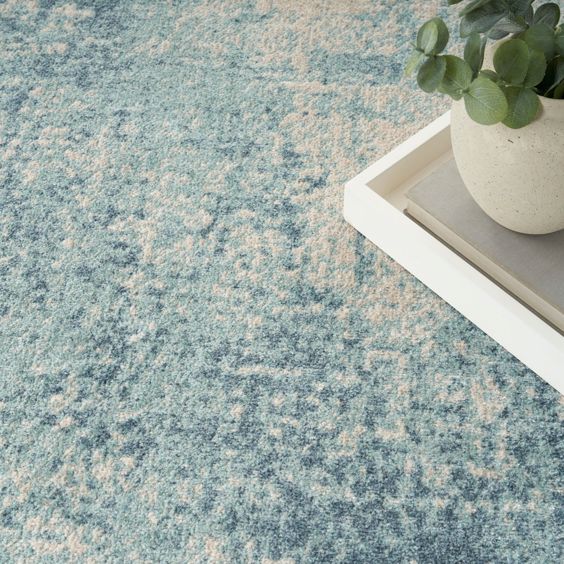 Blue textured rug with beige speckles; white rectangular tray holds a round planter with green eucalyptus atop stacked books.