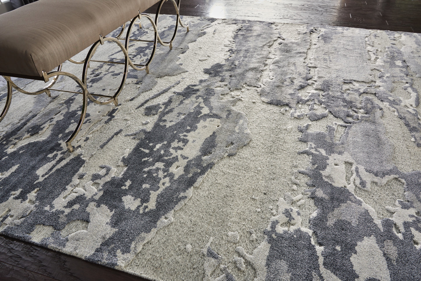 Abstract gray & cream area rug with irregular pattern under a beige upholstered bench with circular gold metal legs.