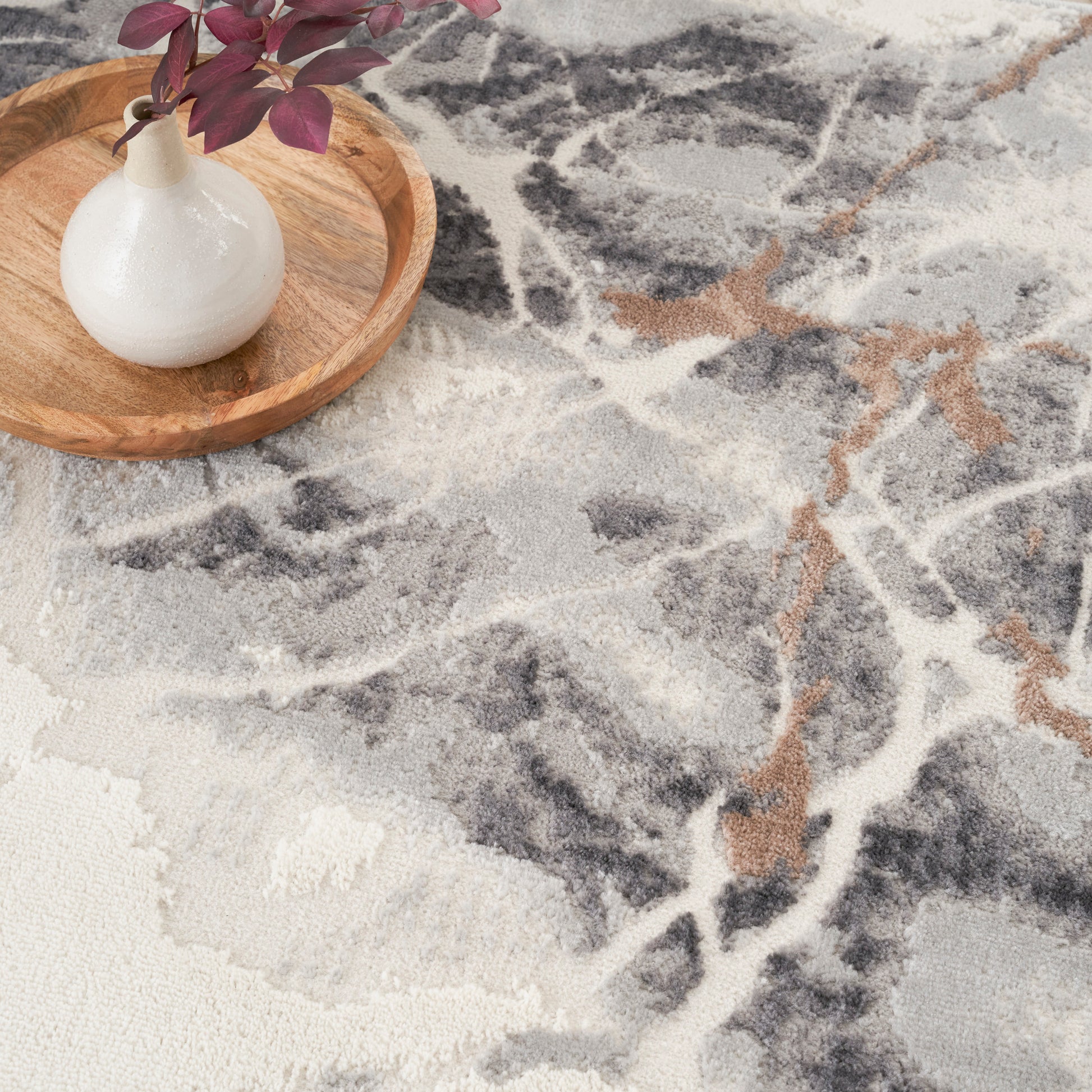Round wooden tray with a white ceramic vase and purple leaves on a grey, white, and tan abstract rug.