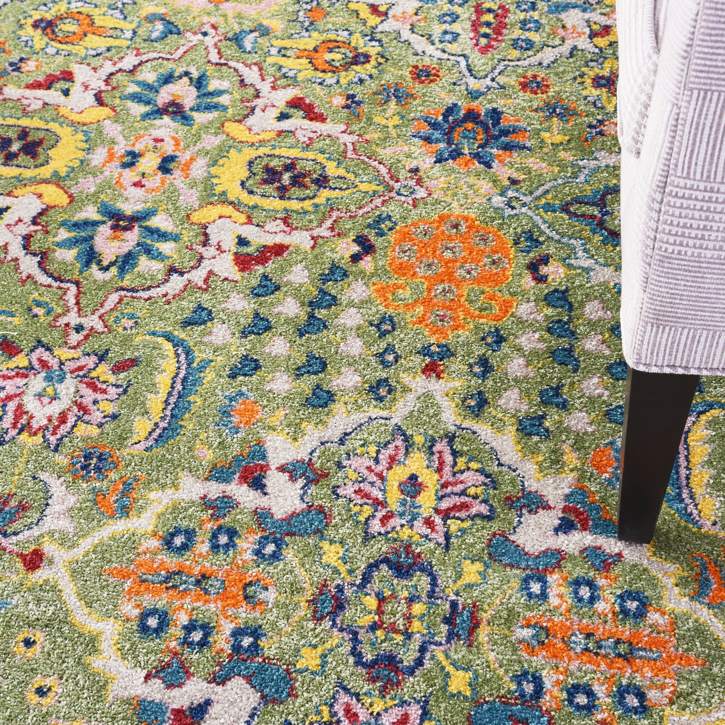 Bright multicolor floral area rug with green background; orange, blue & yellow blossoms; plush pile near a gray sofa leg.