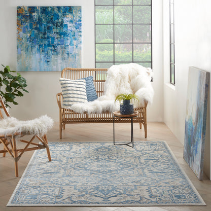 Blue and cream patterned rug in a sunlit living room with wicker seating, faux fur throws, blue cushions, plant, and art.