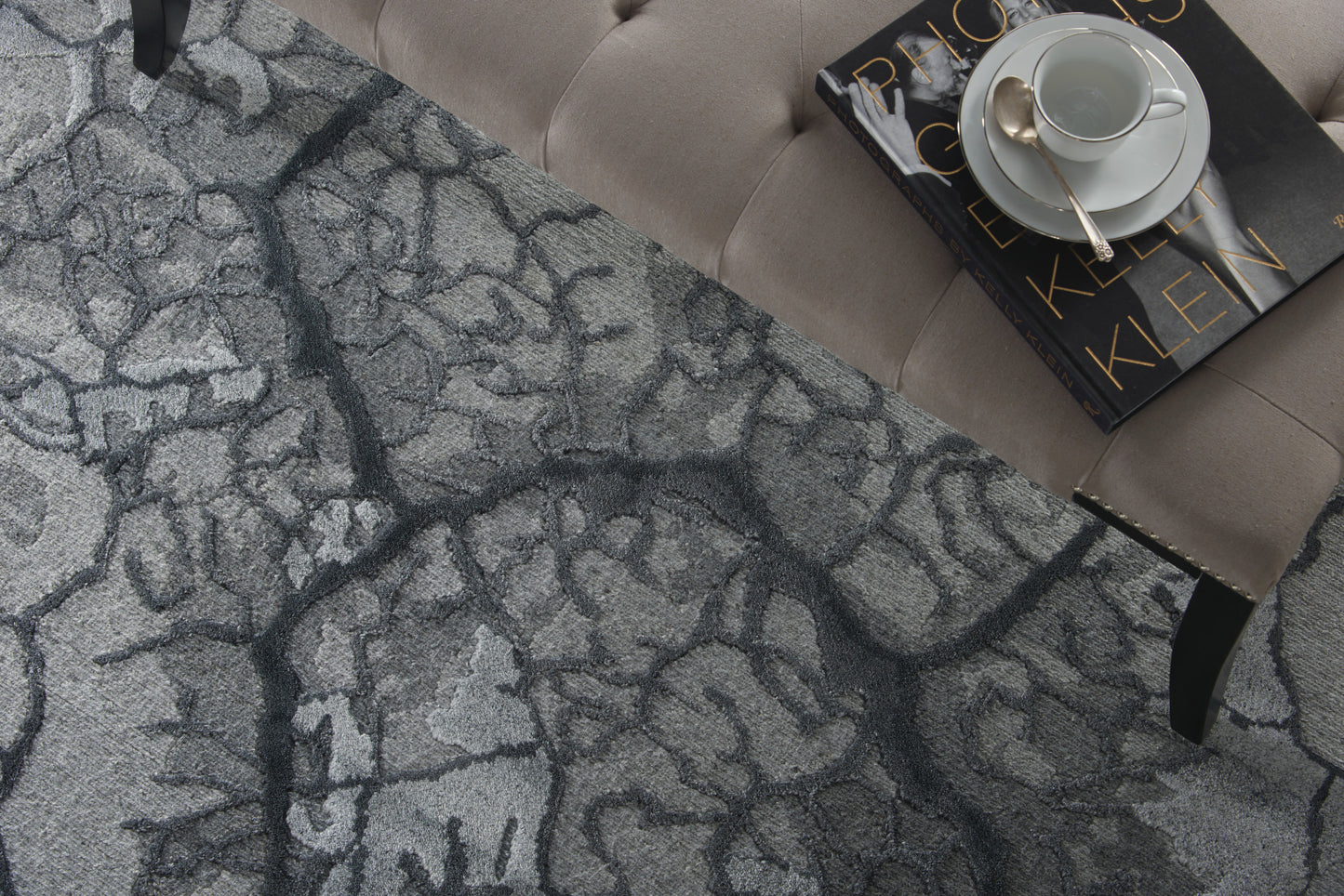 Gray-blue cracked-pattern rug under a beige tufted sofa; white cup and saucer atop a black book with gold lettering.