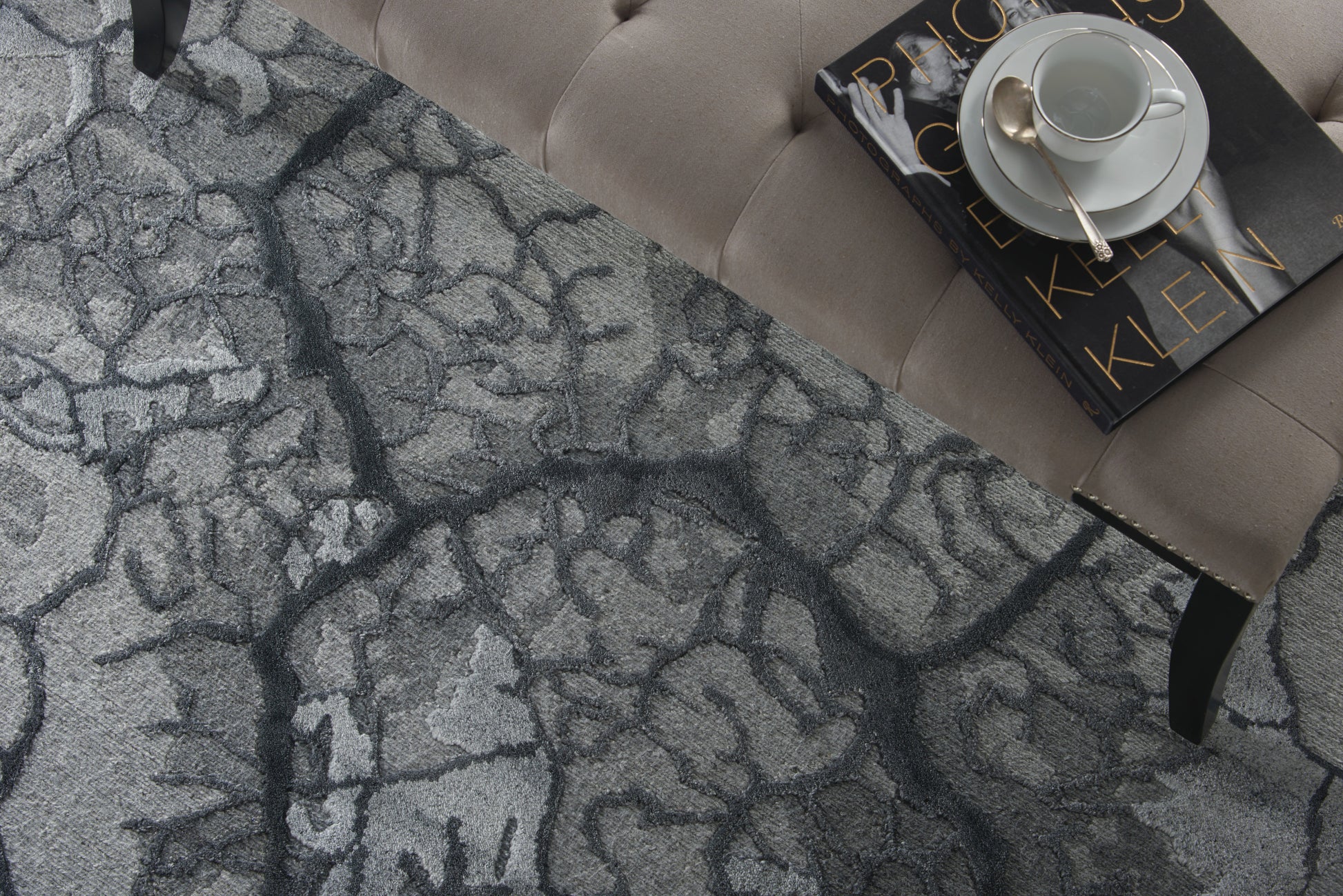 Gray-blue cracked-pattern rug under a beige tufted sofa; white cup and saucer atop a black book with gold lettering.