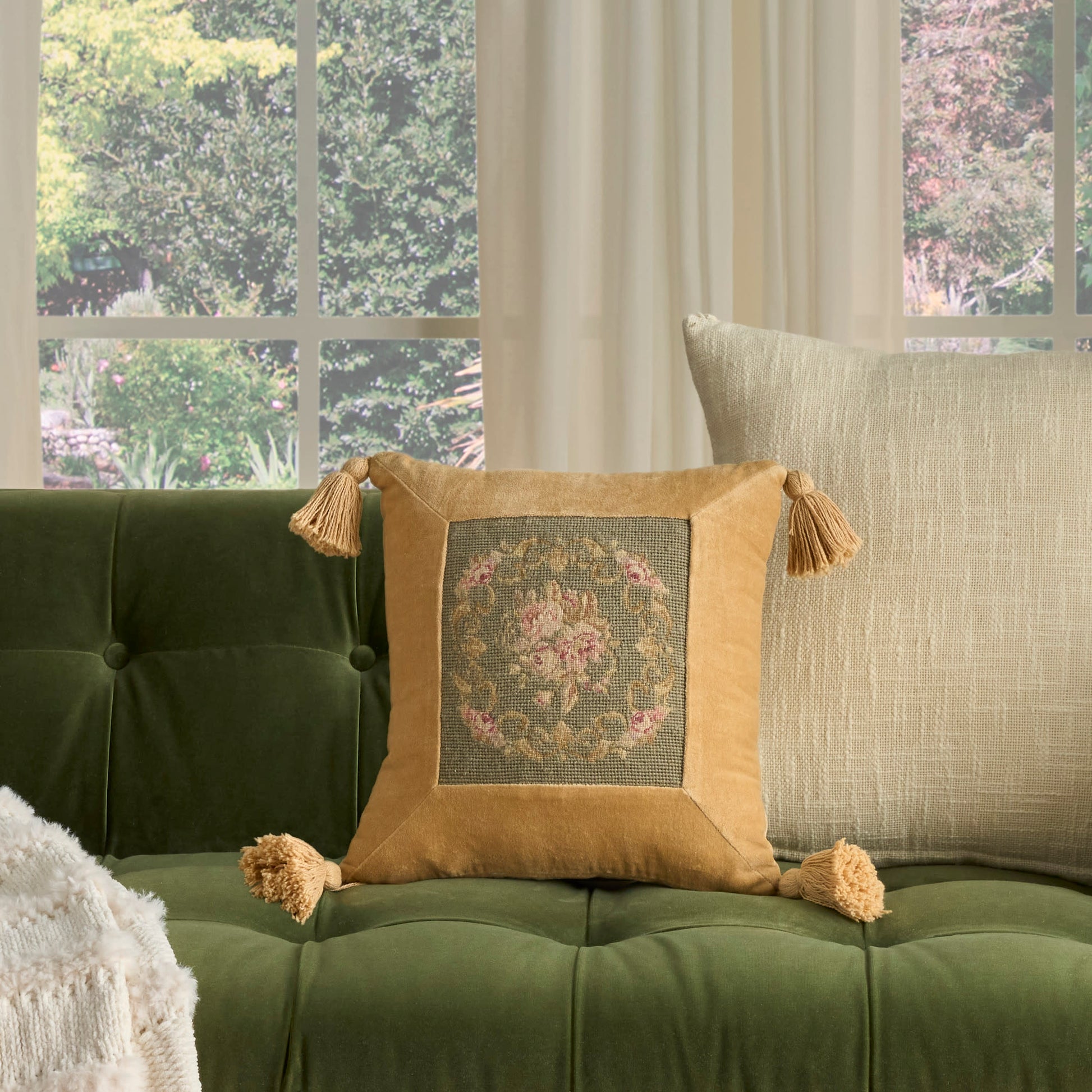 Mustard tassel-trim decorative pillow with floral embroidery on a tufted green velvet sofa, beige back pillow.