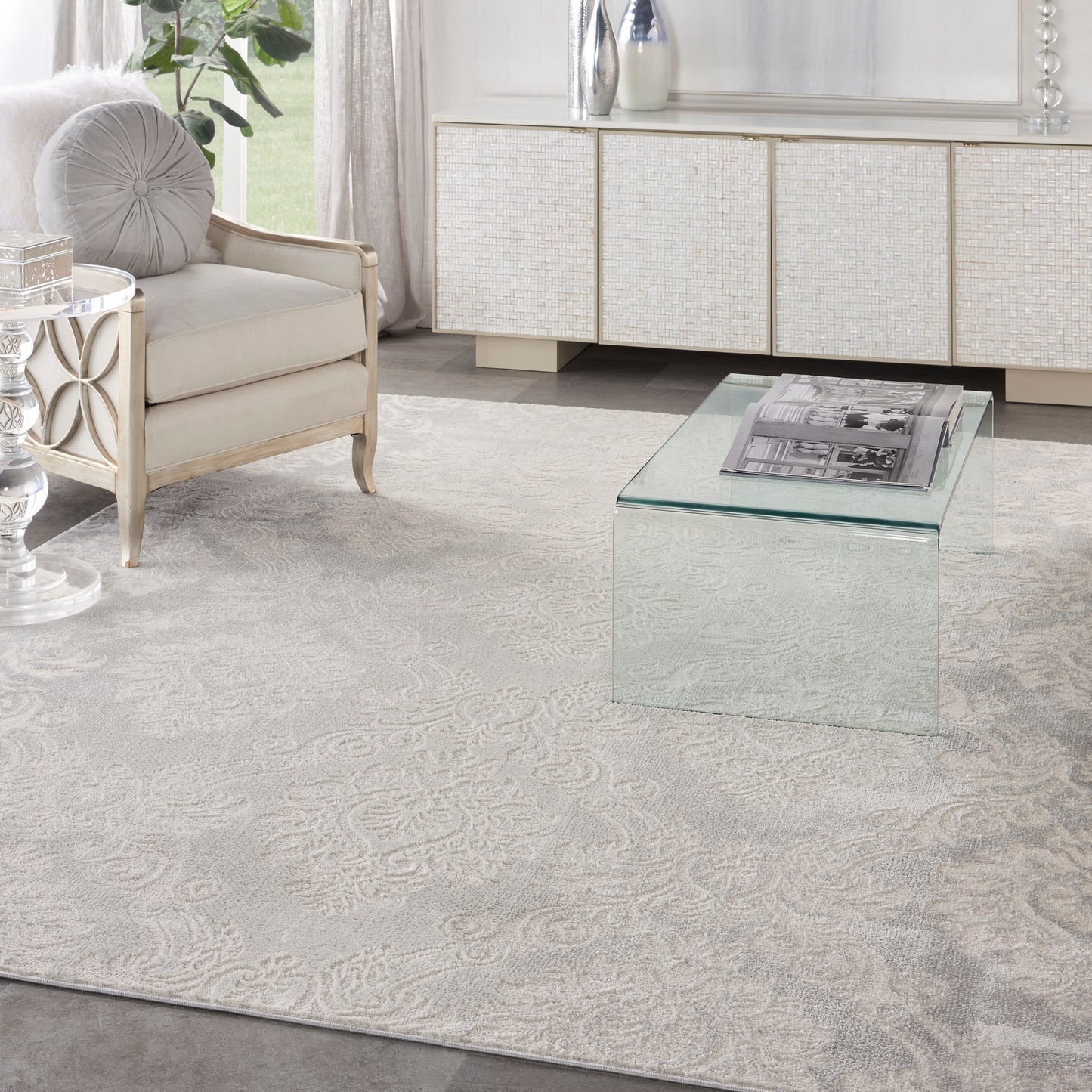 Cream fabric armchair with a round gray cushion beside a glass cube coffee table on a patterned rug in modern living room.