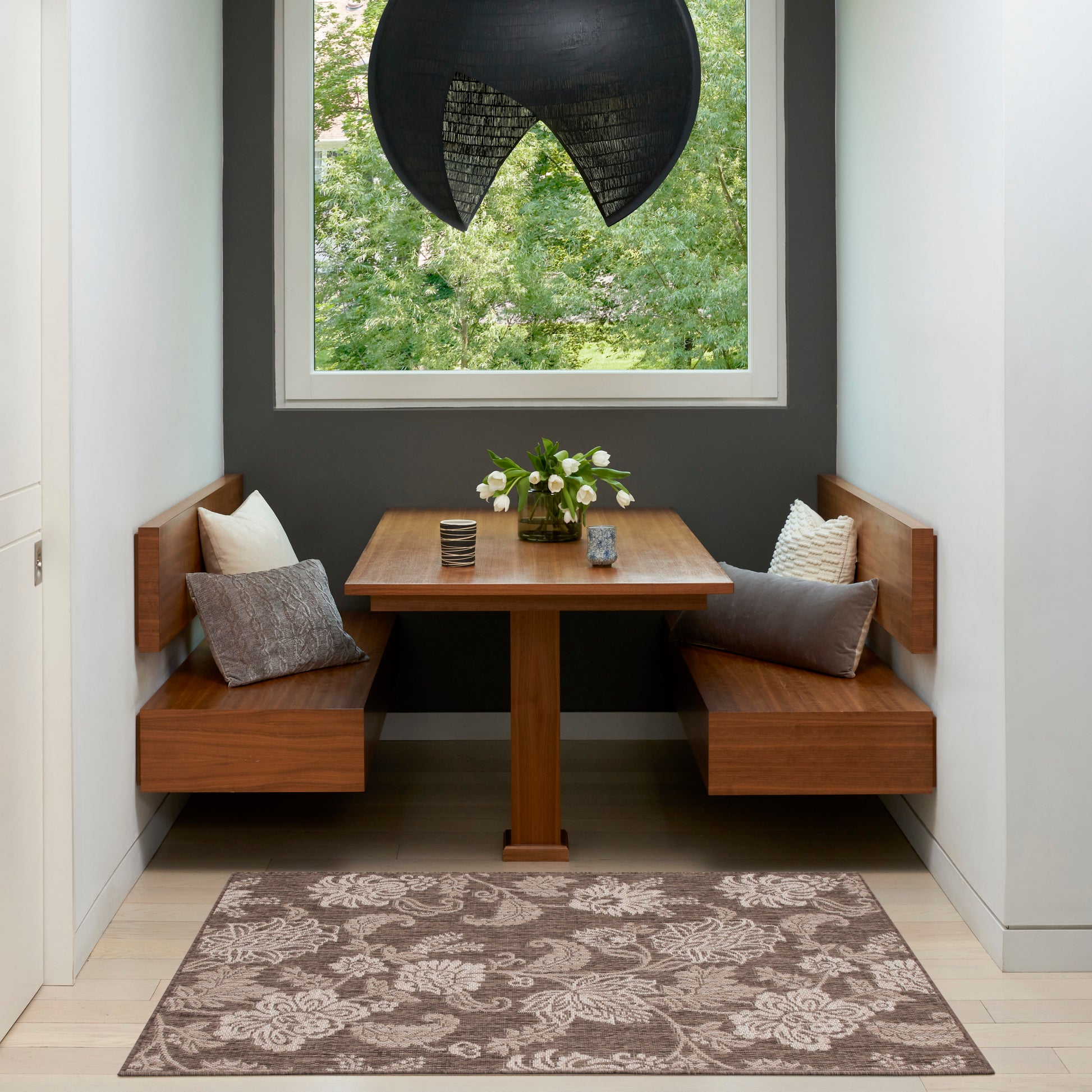Wood corner dining nook with square table, L-shaped benches, grey cushions, vase of white flowers, dark wall, black pendant.