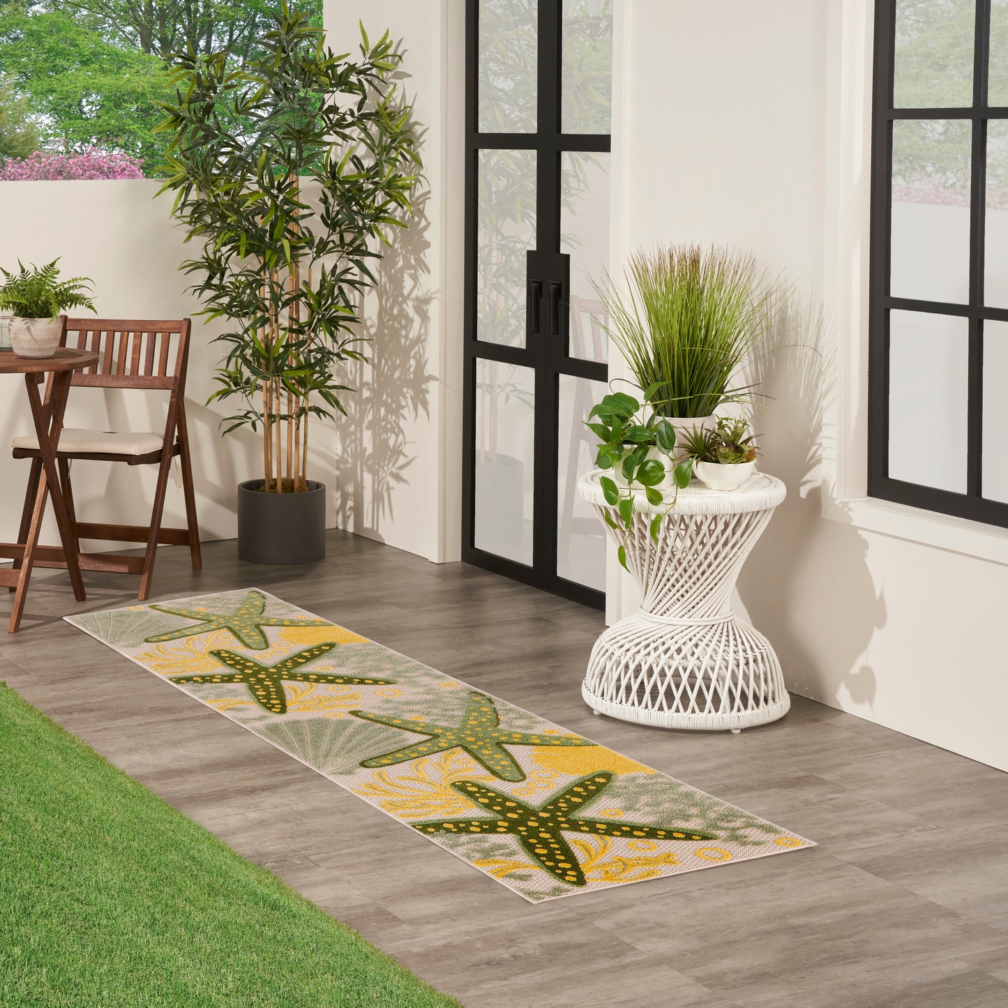 Patio entry with starfish-pattern runner rug in green/yellow, potted plants, wicker table, wooden chair, black-framed doors.