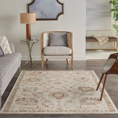 Neutral living room with a light ornate rug, beige upholstered armchair with wooden legs, and a round side table & lamp.