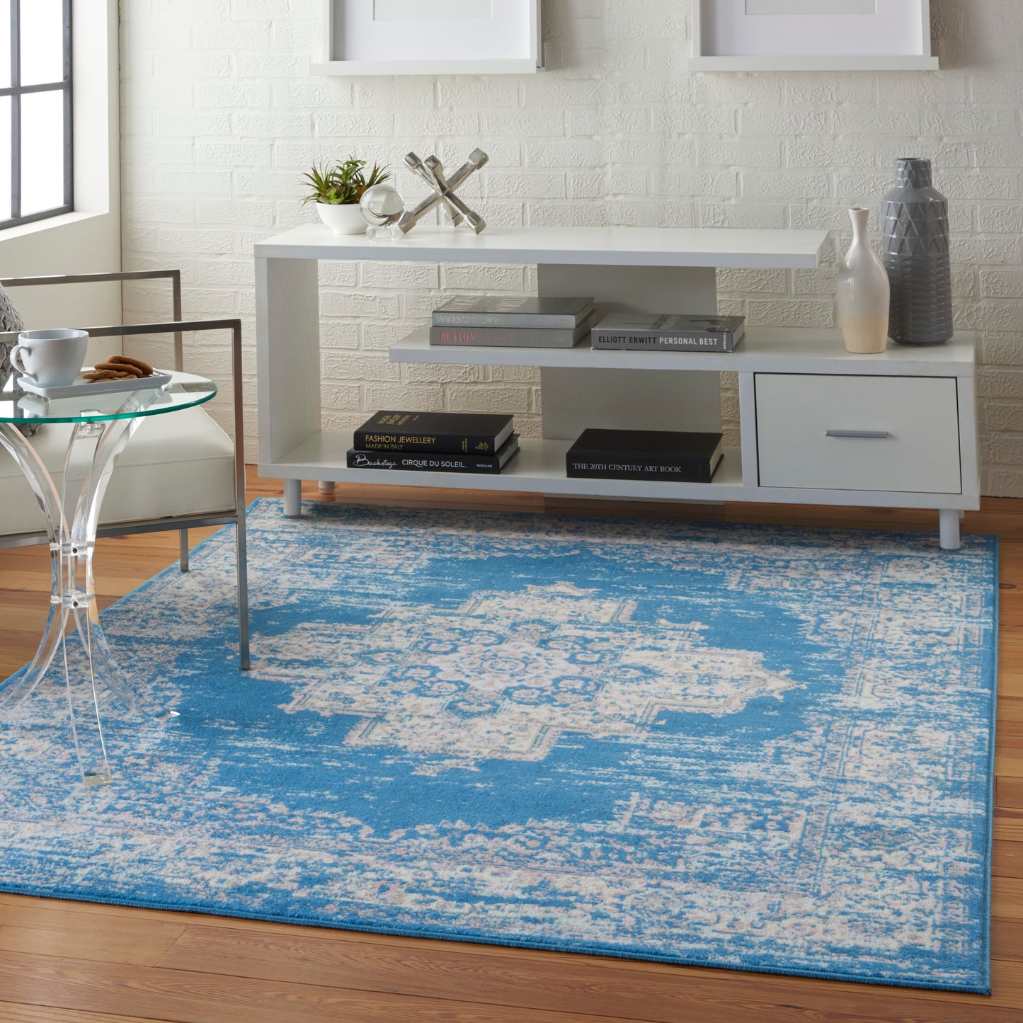 White modern console table with shelves and a drawer, blue distressed rug, glass side table, plant and decorative vases.
