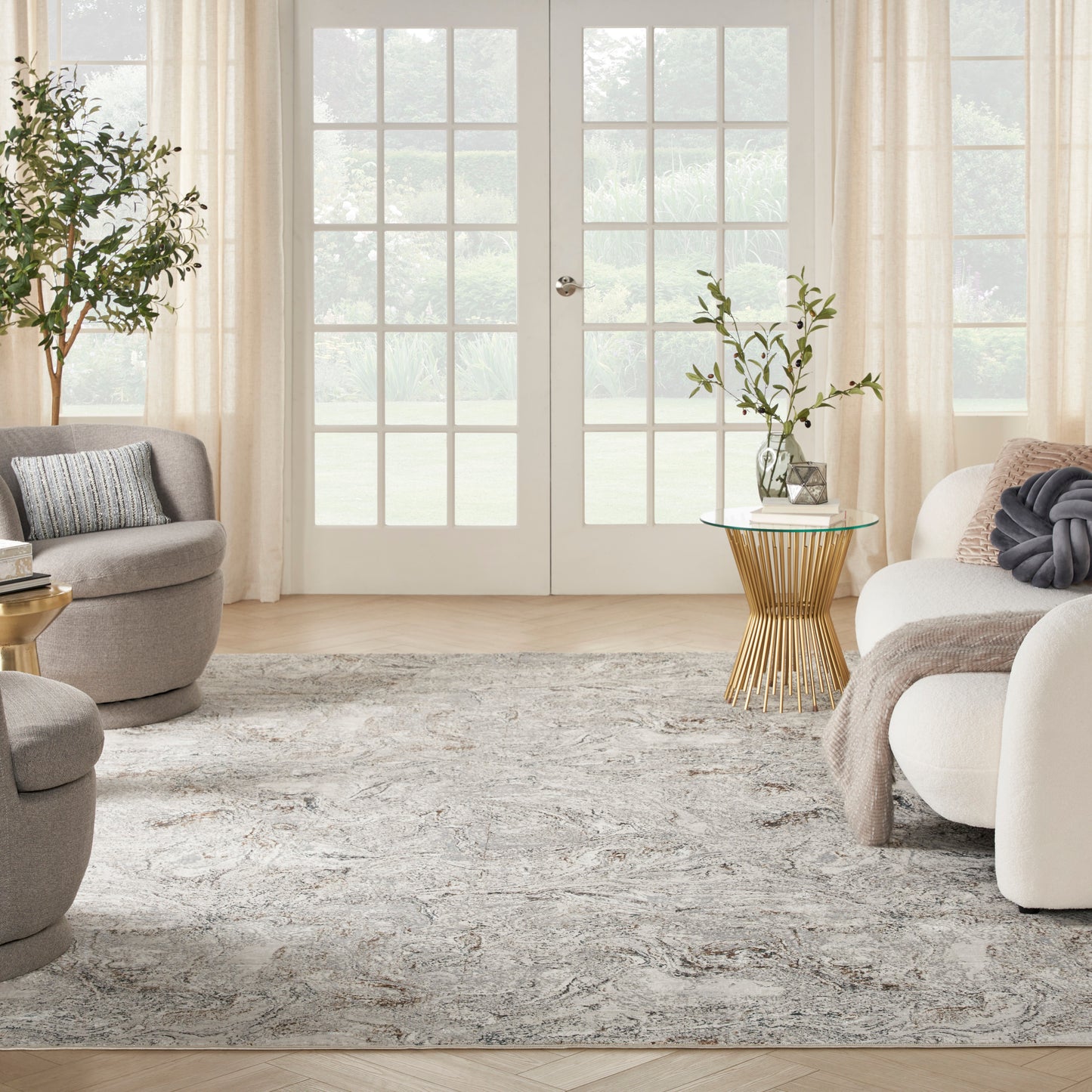 Bright contemporary living room with French doors, beige drapes, gray rounded chairs, white sofa, textured rug, gold metal side table with plant.