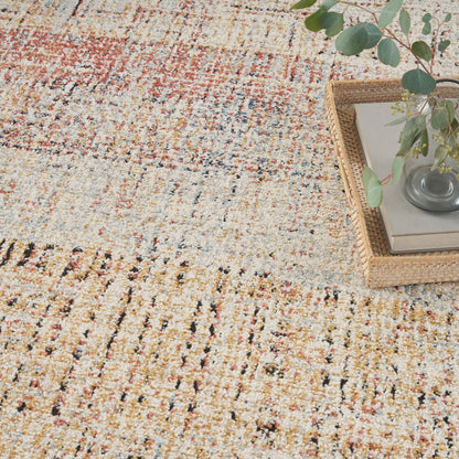 Neutral woven area rug with rust-orange and blue specks; corner wicker tray holding books and a glass vase of eucalyptus.