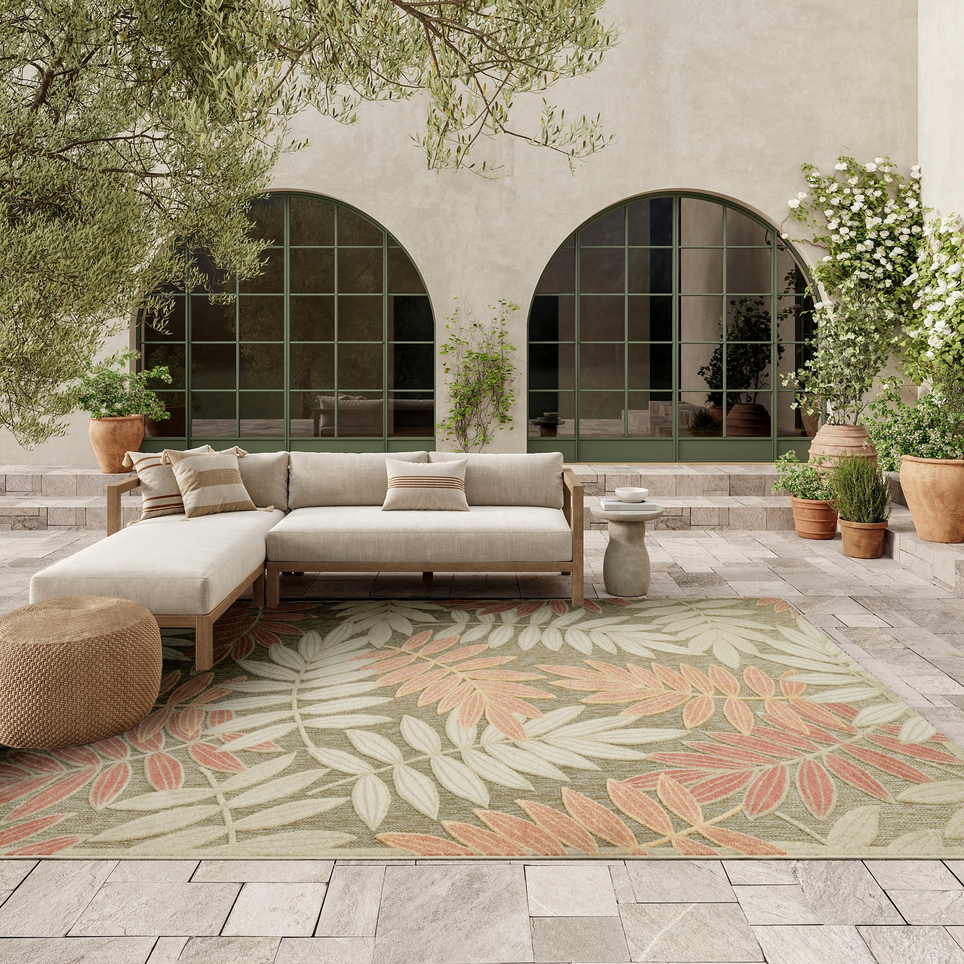 Outdoor patio with beige fabric sectional on a wooden frame, palm-leaf rug, woven pouf, terracotta pots & arched windows.