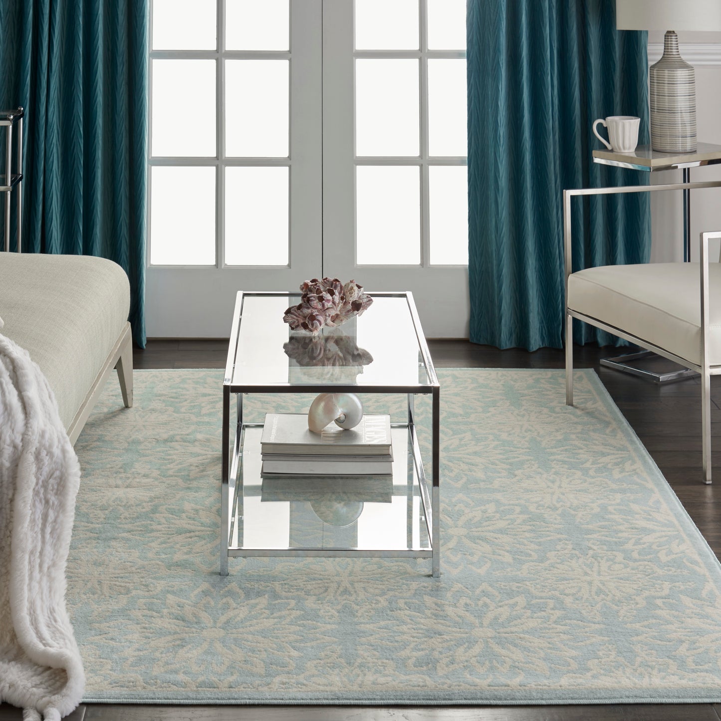 Patterned pale blue rug on the floor of a living room with teal curtains, white sofa & seashell decor.