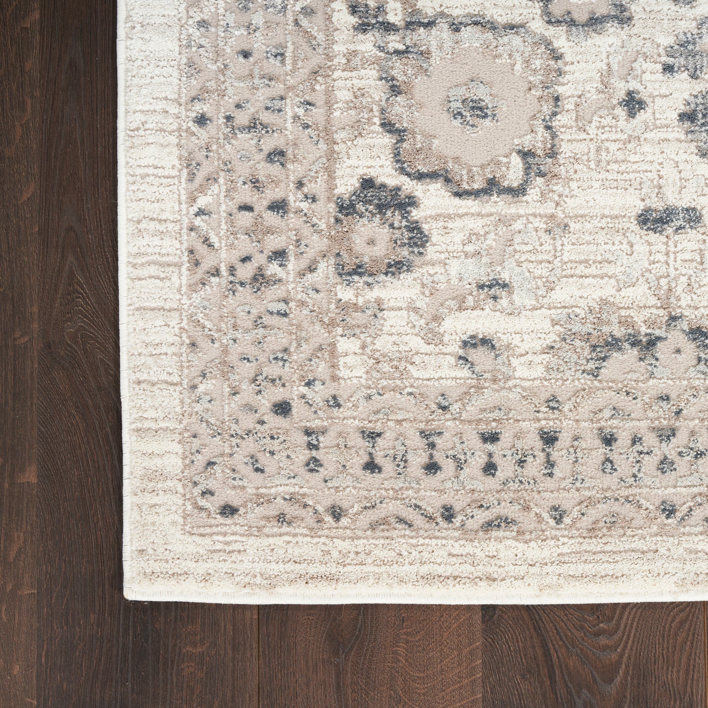 Beige oriental rug with floral border and muted blue-gray motifs on dark wood floor; close-up corner.