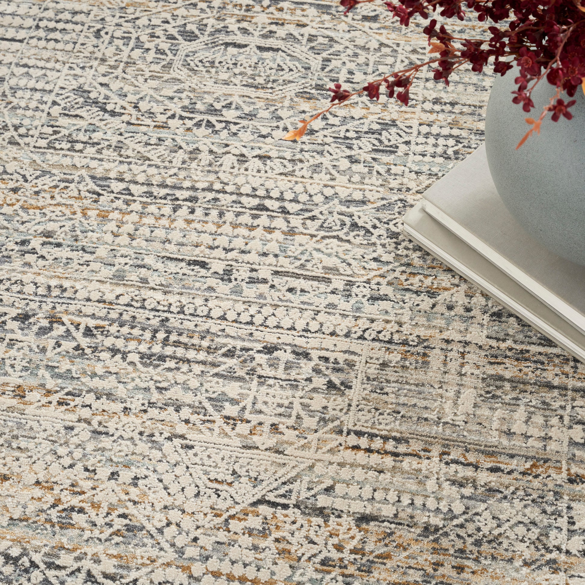 Muted gray-blue patterned area rug with cream and tan accents; vase of red dried flowers atop stacked books.