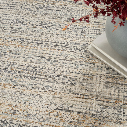 Muted gray-blue patterned area rug with cream and tan accents; vase of red dried flowers atop stacked books.