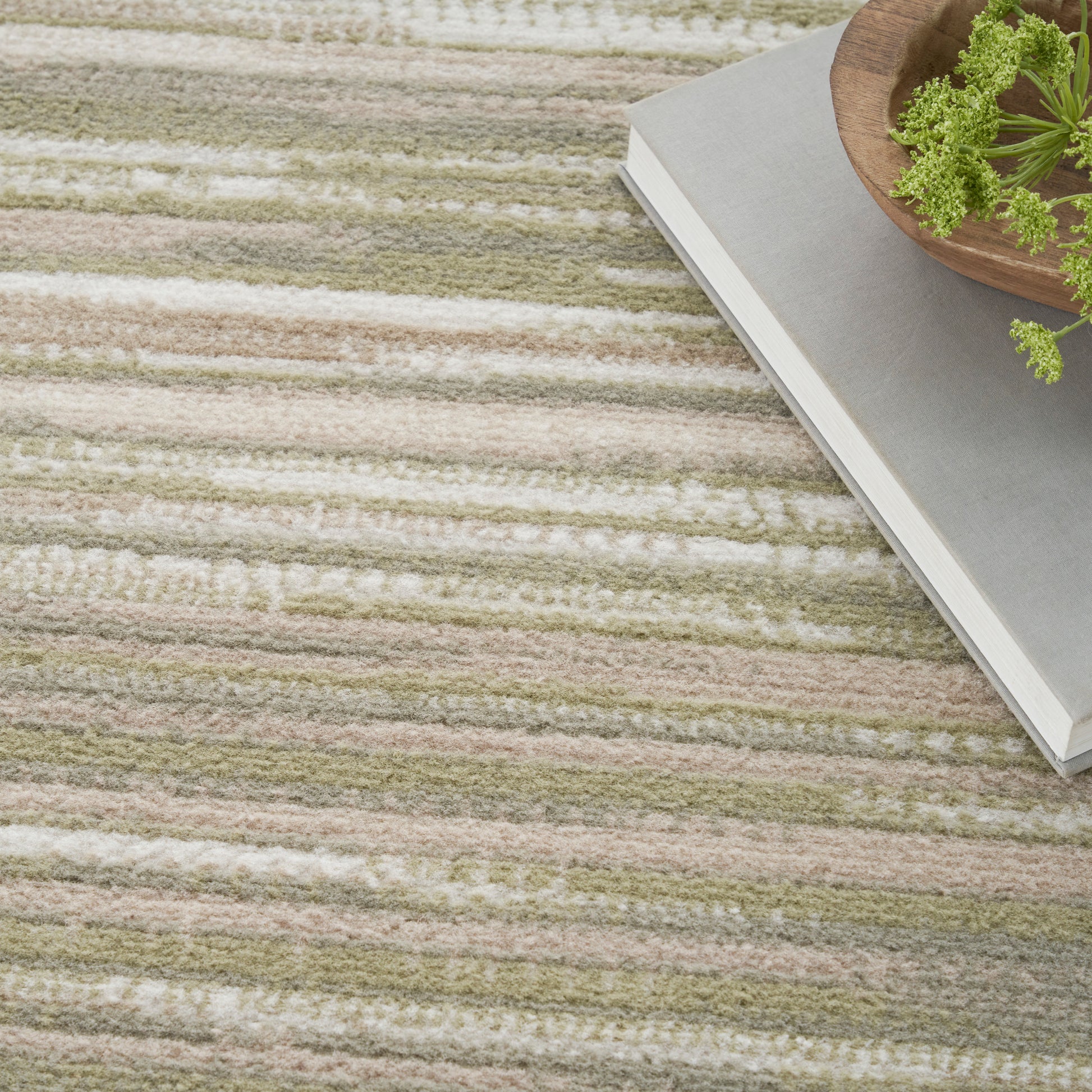 Striped beige, cream and olive rug with soft pile, topped by a gray book and a wooden bowl with greenery.