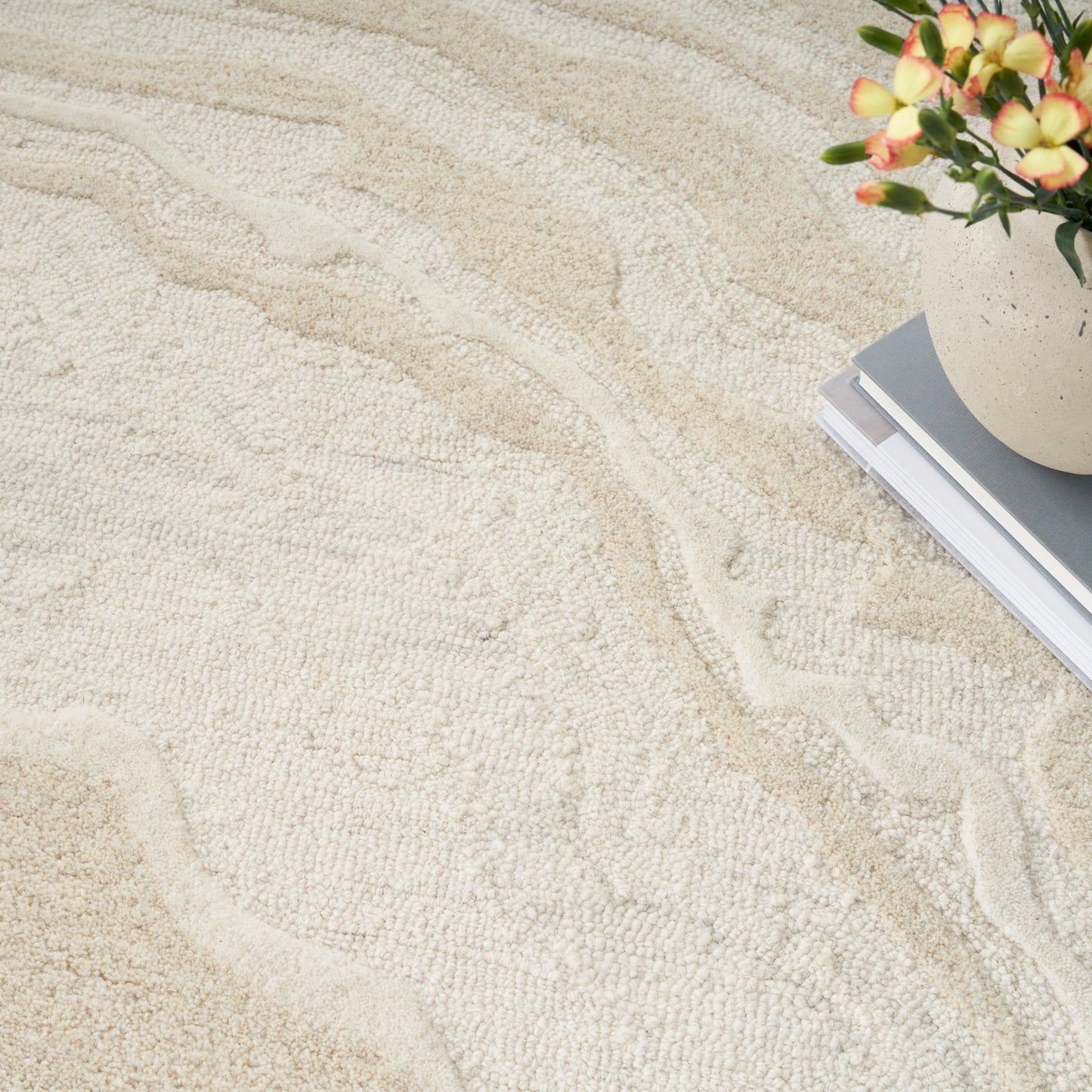 Beige textured rug with a wave pattern; top-right shows a flower pot and a stack of magazines.