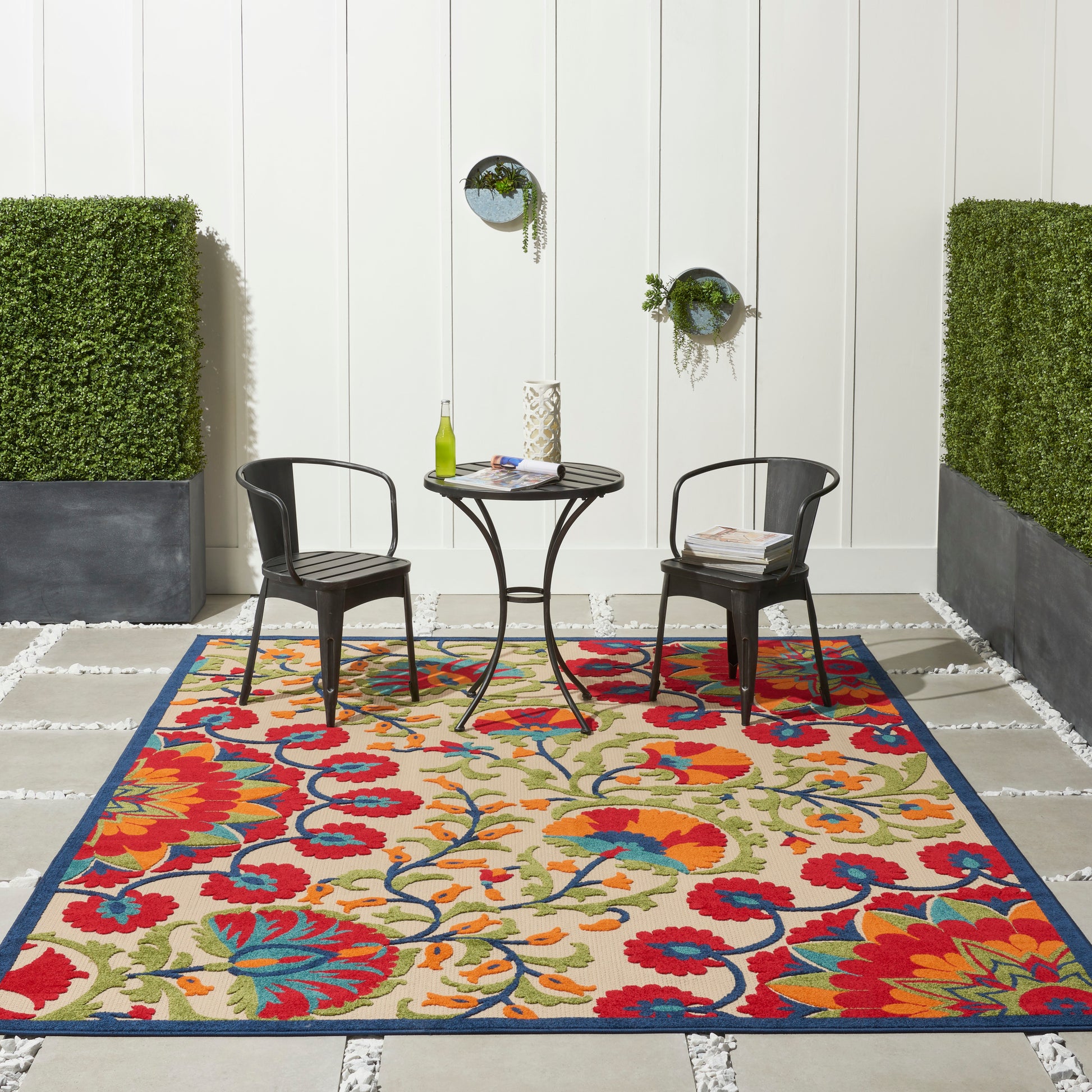 Outdoor seating area with a colorful floral rug, two black metal chairs and a round table against a white paneled wall.