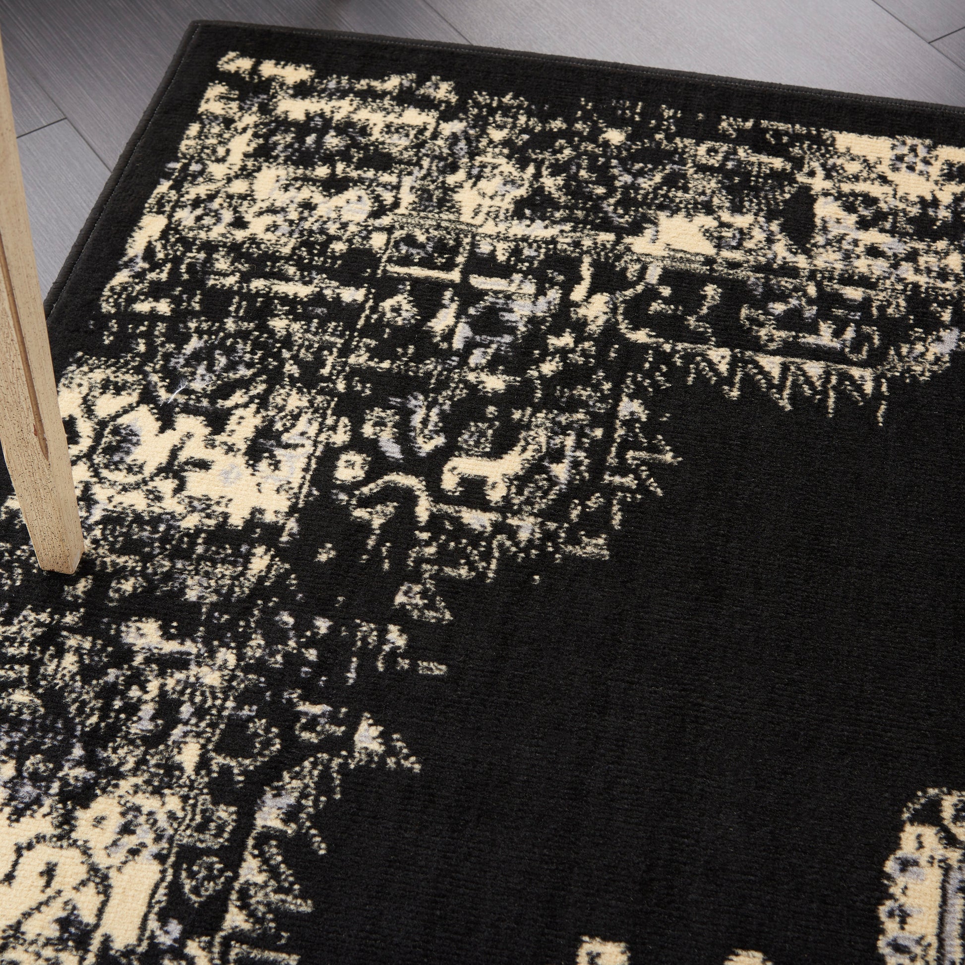 Rectangular black and ivory distressed-pattern rug with abstract floral motifs and a wooden table leg on gray floor.