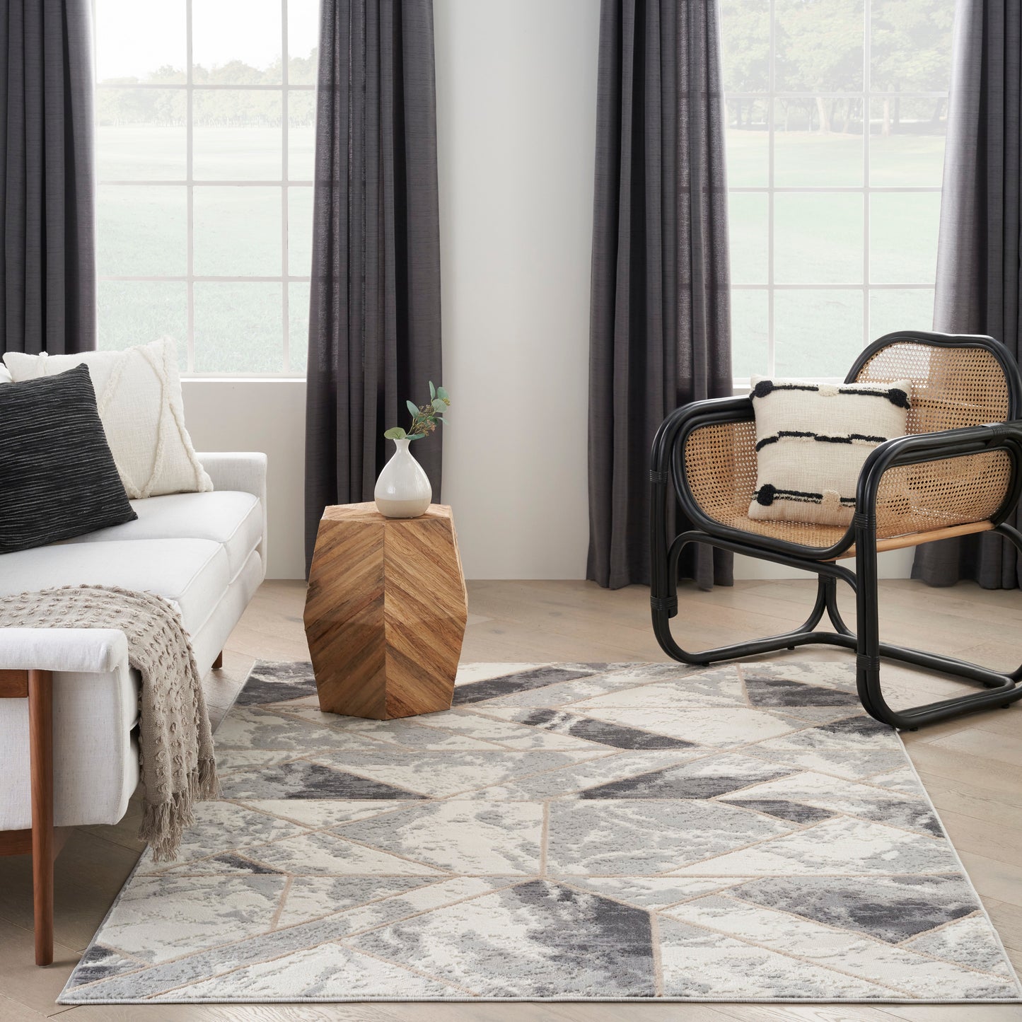 Modern living room with white sofa, gray geometric rug, wooden side table with vase, and a black-framed rattan lounge chair.