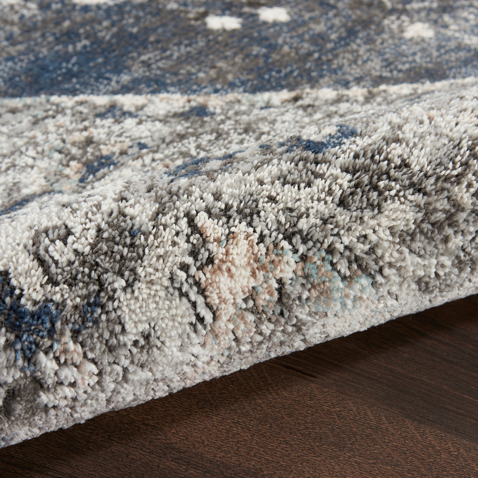 Close-up of a plush shag rug with blue, gray, and ivory tones and a thick, dense pile on a dark wood floor.