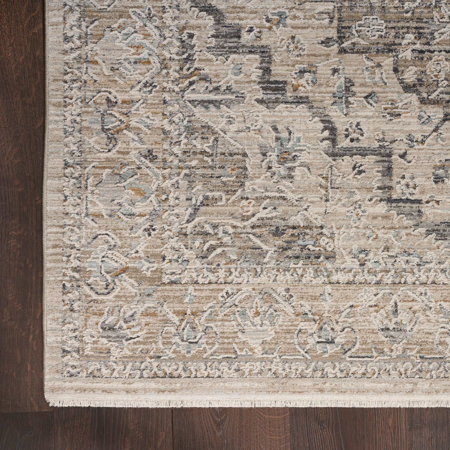 Distressed beige and gray oriental rug with blue accents, ornate floral border and fringe on dark wood floor