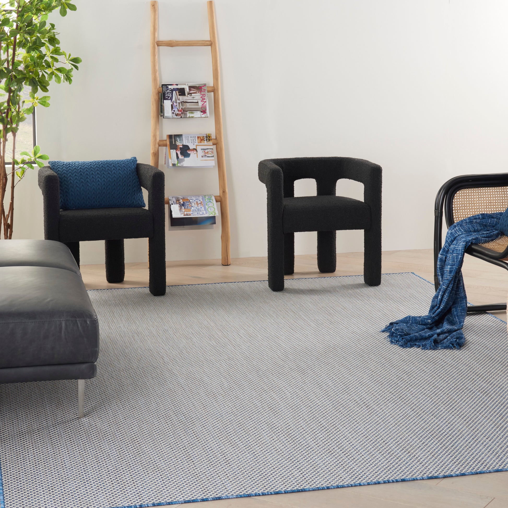 Modern living room with black sculptural armchairs, gray rug, wooden ladder shelf, leafy plant, and a blue throw blanket.
